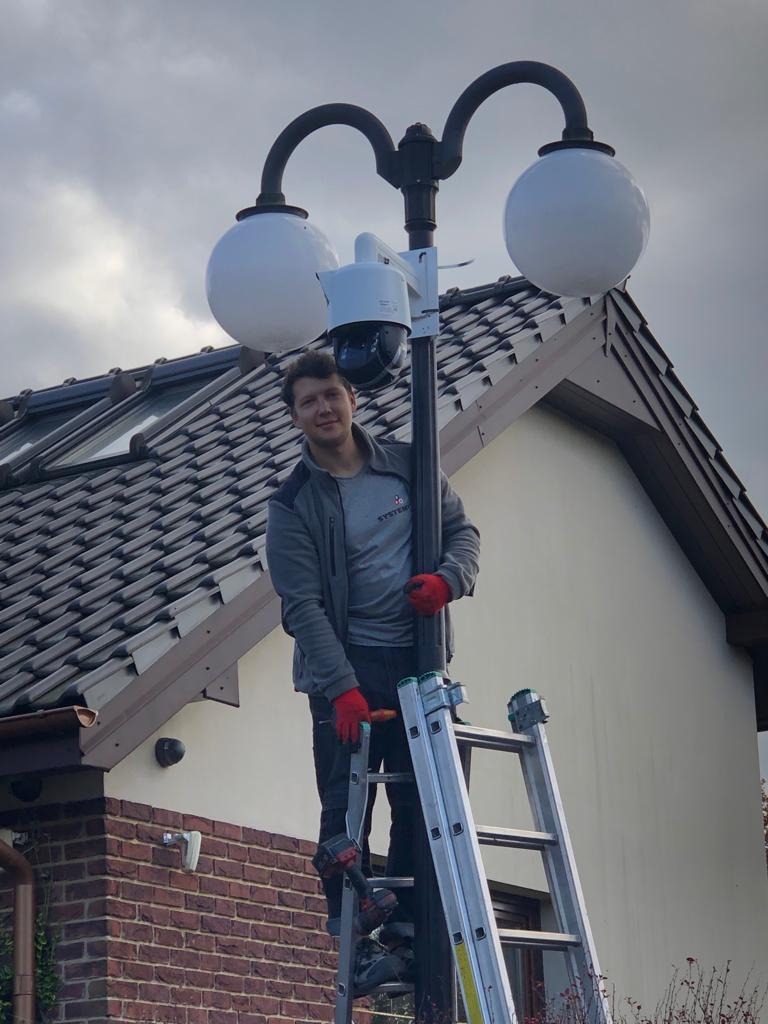 Mężczyzna na drabinie instaluje kamerę monitoringu na słupie oświetleniowym z dwoma kulistymi kloszami, widoczny budynek z dachówką.