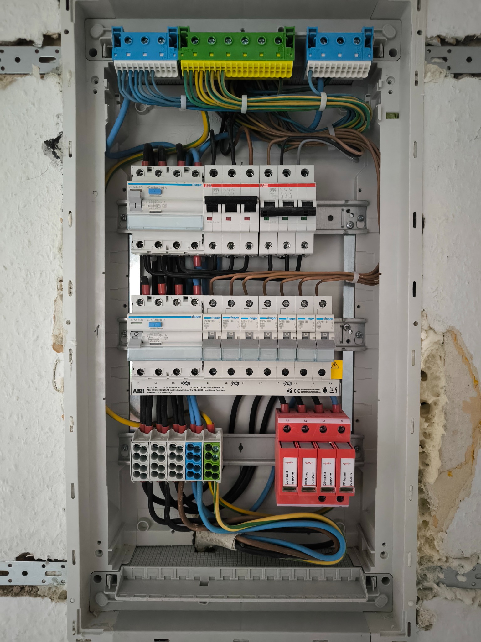 Otwarta skrzynka elektryczna z zainstalowanymi bezpiecznikami, rozłącznikami, ogranicznikami przepięć i listwami zaciskowymi, widoczne okablowanie, ściana z widocznymi śladami po naprawach.