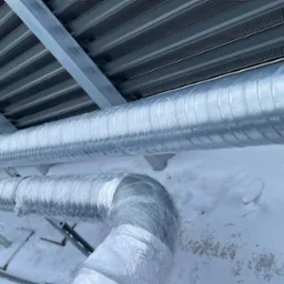Dwie izolowane rury wentylacyjne zamontowane na zewnątrz budynku pokrytego śniegiem, widoczne metalowe wsporniki i elementy konstrukcyjne dachu.