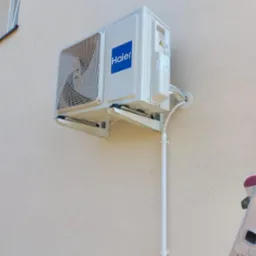 Biały klimatyzator Haier zamontowany na beżowej ścianie budynku, widoczna drabina aluminiowa z naklejkami.