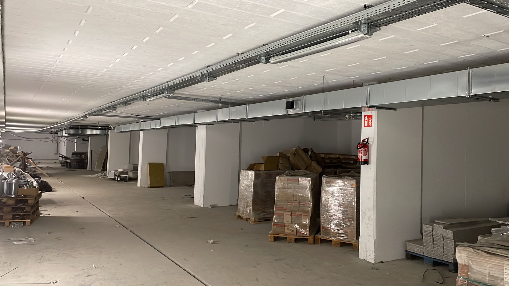 Wnętrze budynku komercyjnego w trakcie instalacji systemu wentylacyjnego: widoczne kanały wentylacyjne pod sufitem, palety z materiałami budowlanymi i gaśnica na filarze.