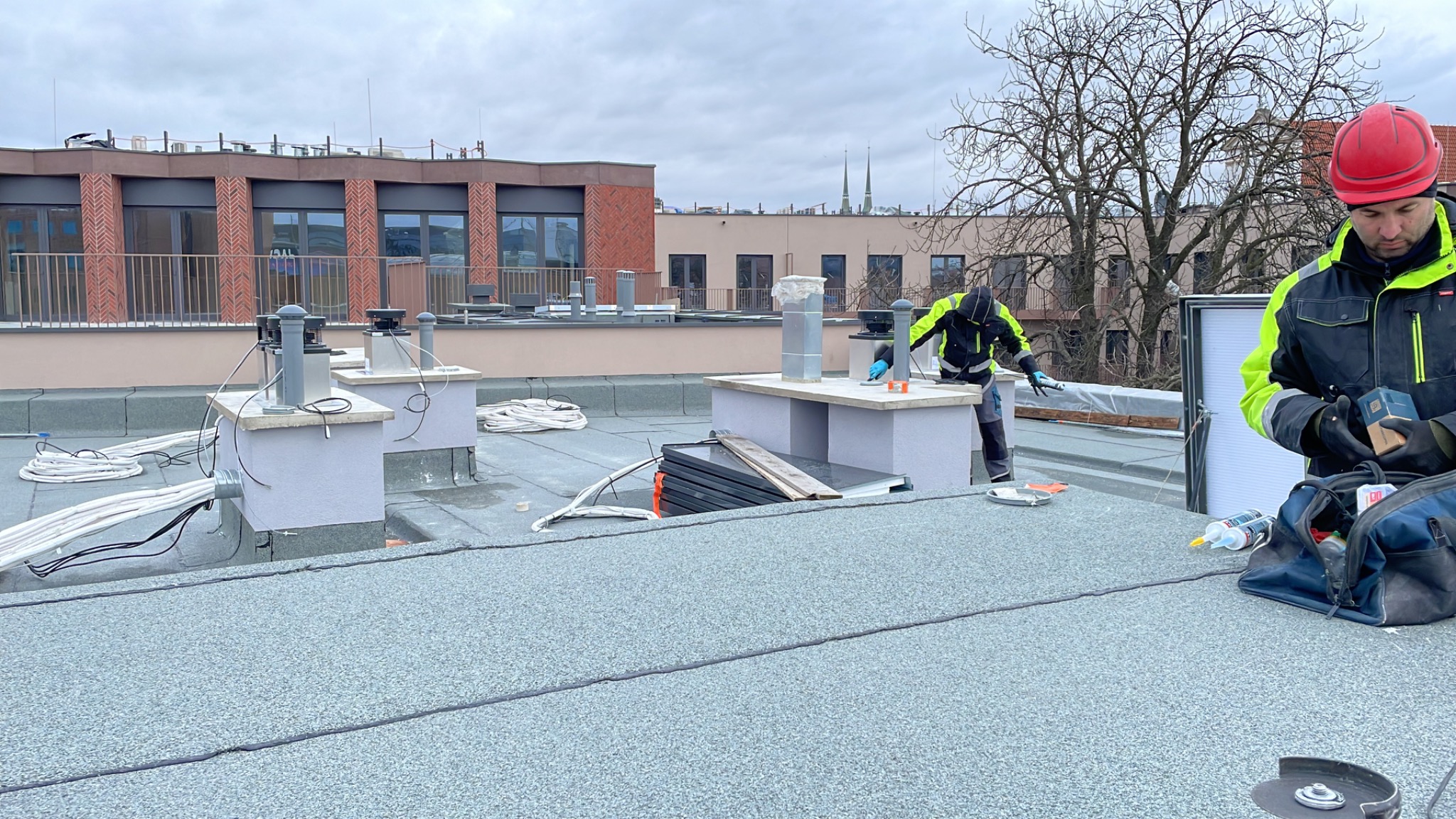 Praca na dachu: Dwóch monterów w odblaskowych kurtkach instaluje system wentylacji z elementami rekuperacji na płaskim dachu budynku. Widoczne przewody, narzędzia i elementy wentylacyjne.