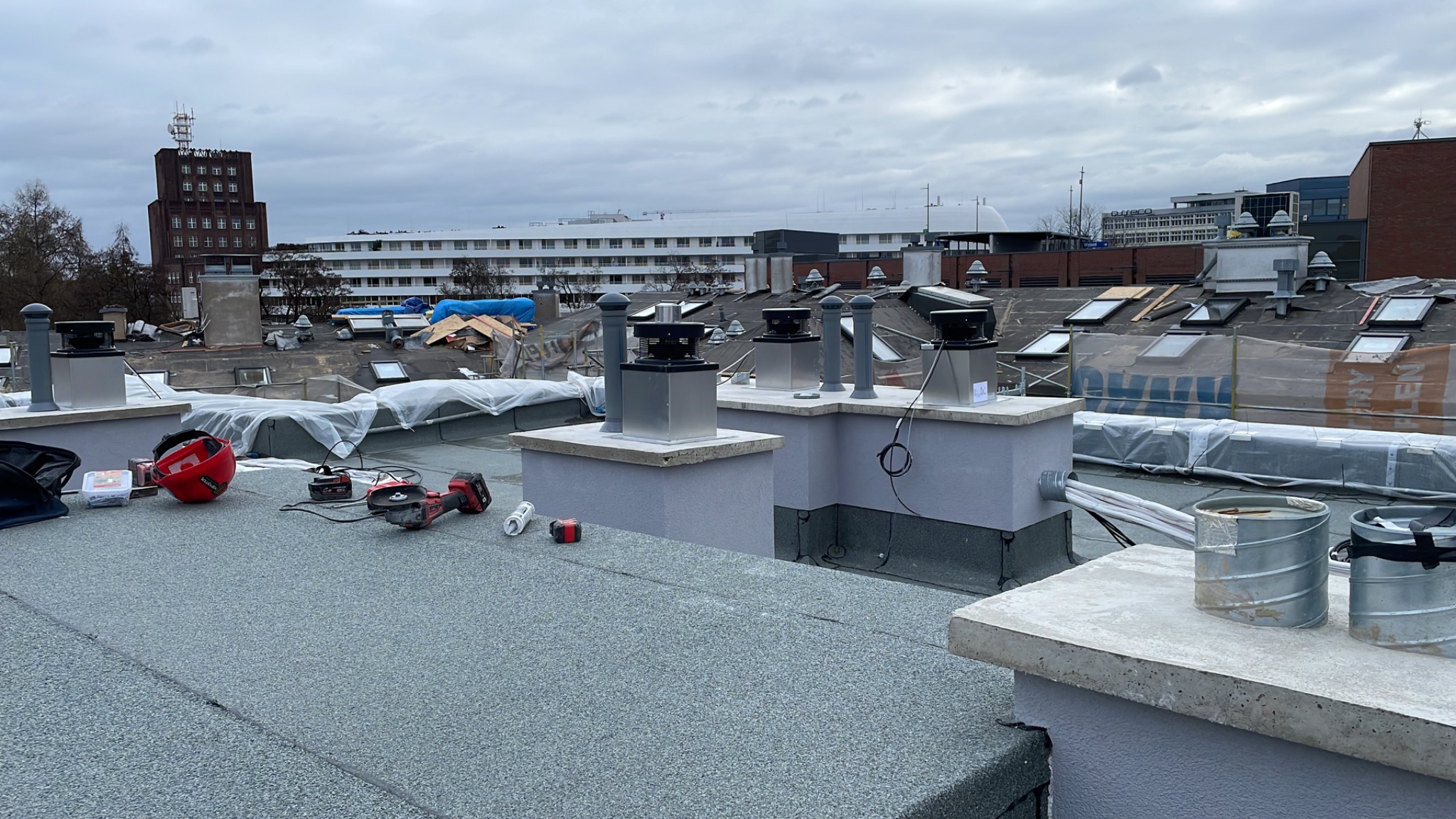 Instalacja systemów wentylacyjnych na dachu budynku, widoczne elementy wentylacyjne, narzędzia pracy, kask ochronny i metalowe kanały wentylacyjne. W tle zabudowa miejska.