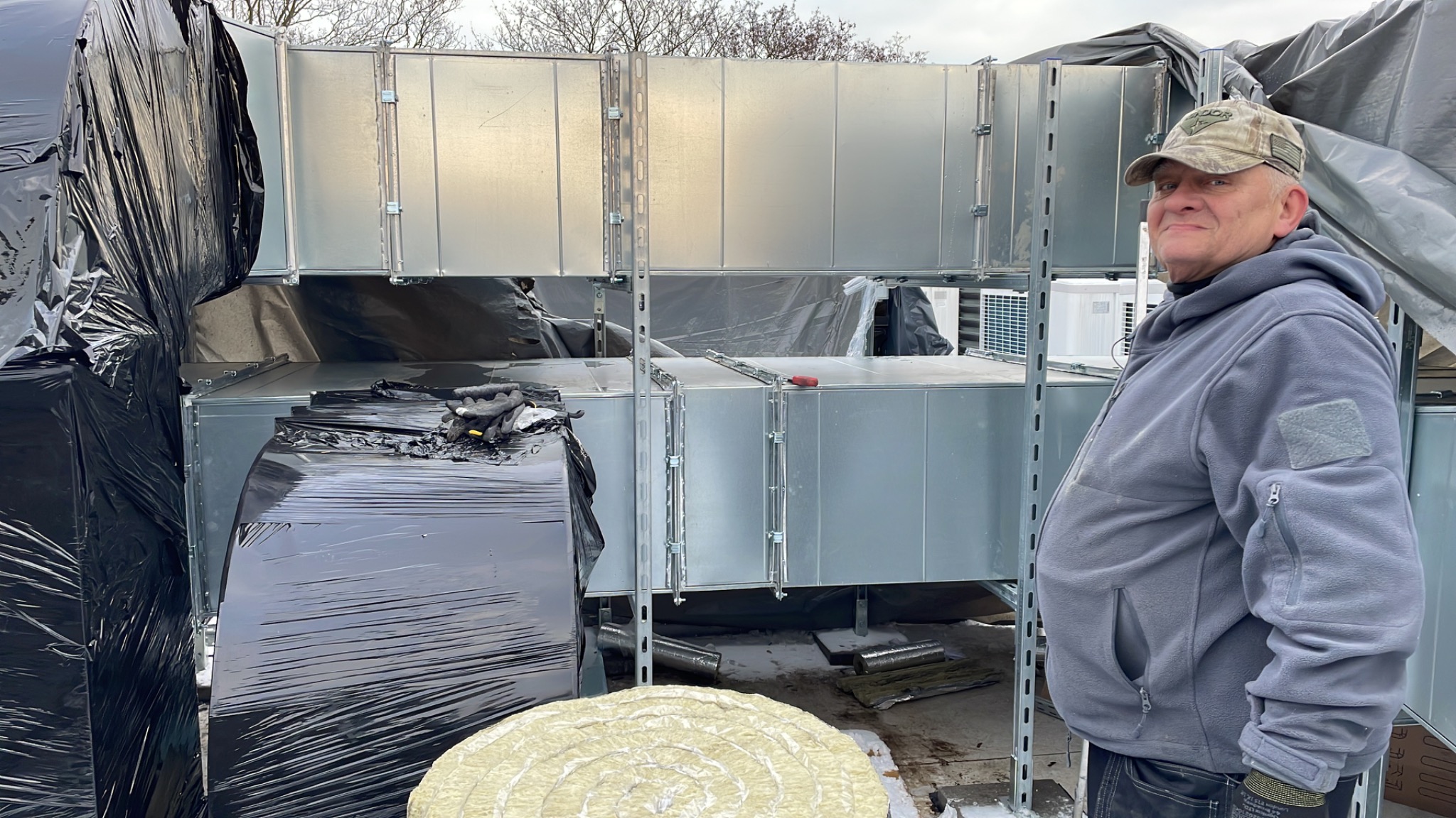 Instalacja kanałów wentylacyjnych z uśmiechniętym instalatorem w czapce moro, widoczne izolowane rury i elementy montażowe.