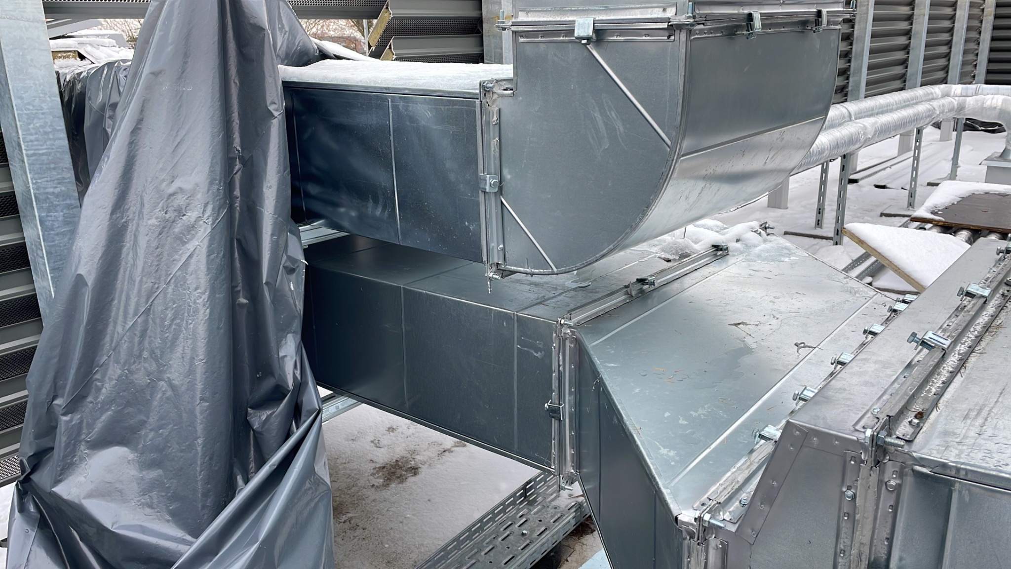 Fragment instalacji wentylacyjnej z metalowych kanałów, częściowo przykrytej folią ochronną na zewnątrz budynku w zimowych warunkach.