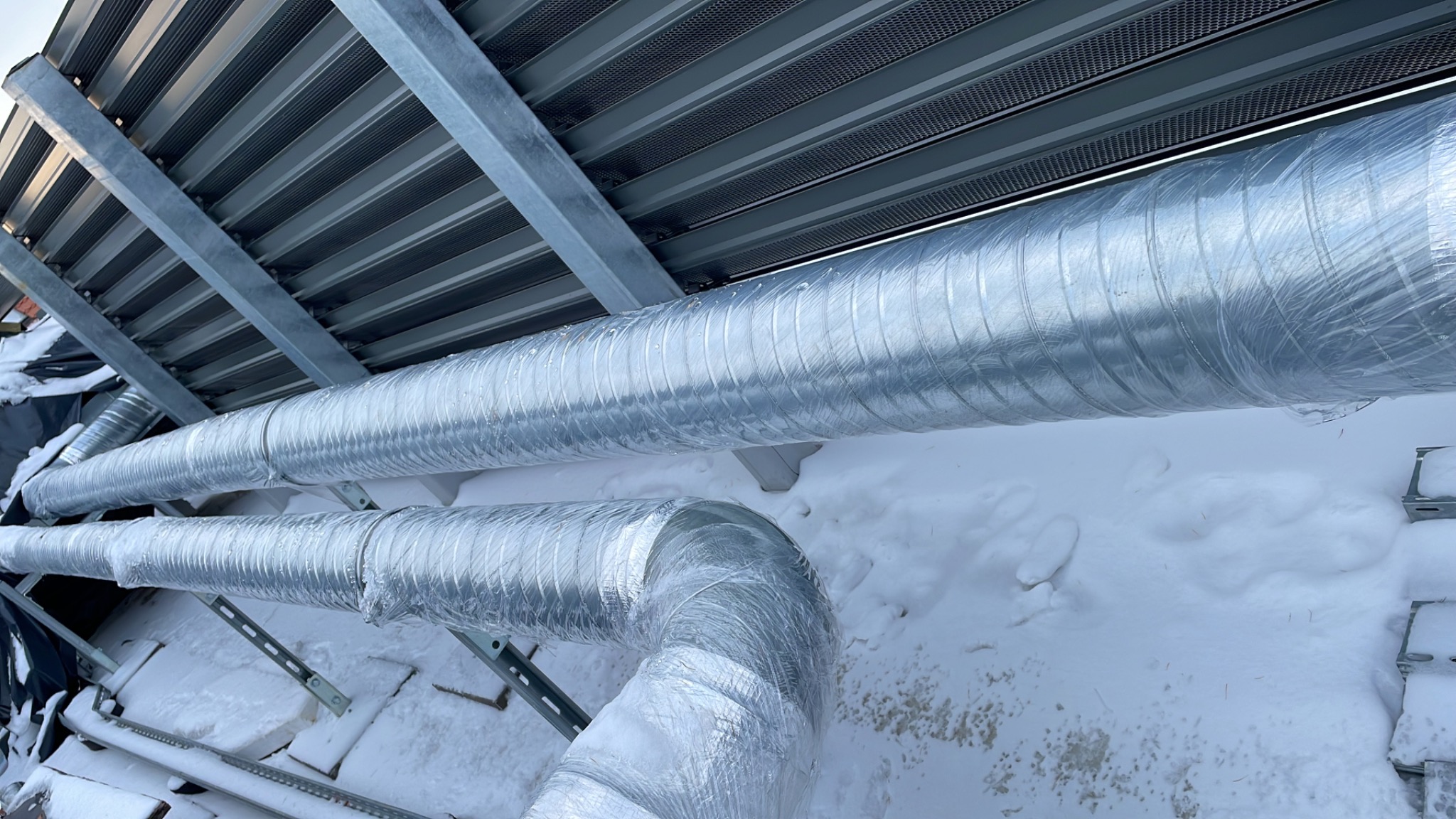 Dwie izolowane rury wentylacyjne zamontowane na zewnątrz budynku pokrytego śniegiem, widoczne metalowe wsporniki i elementy konstrukcyjne dachu.