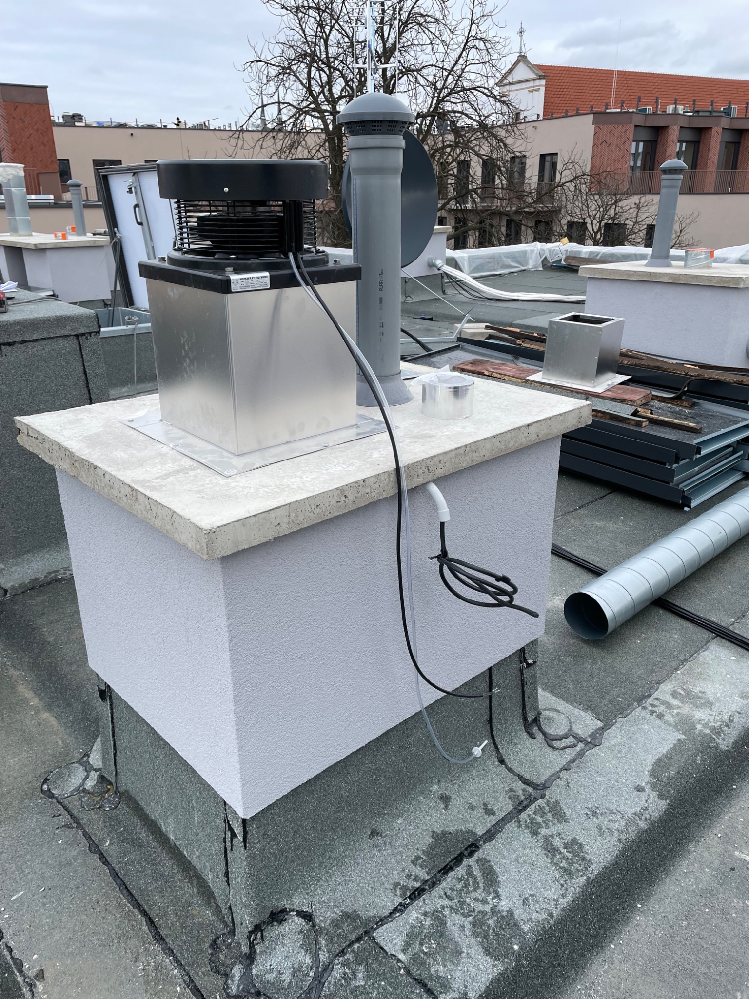 Montaż rekuperacji na dachu budynku: widoczny centralny element rekuperatora z metalową obudową, rurami i przewodami, elementy wentylacyjne obok, dach pokryty papą.