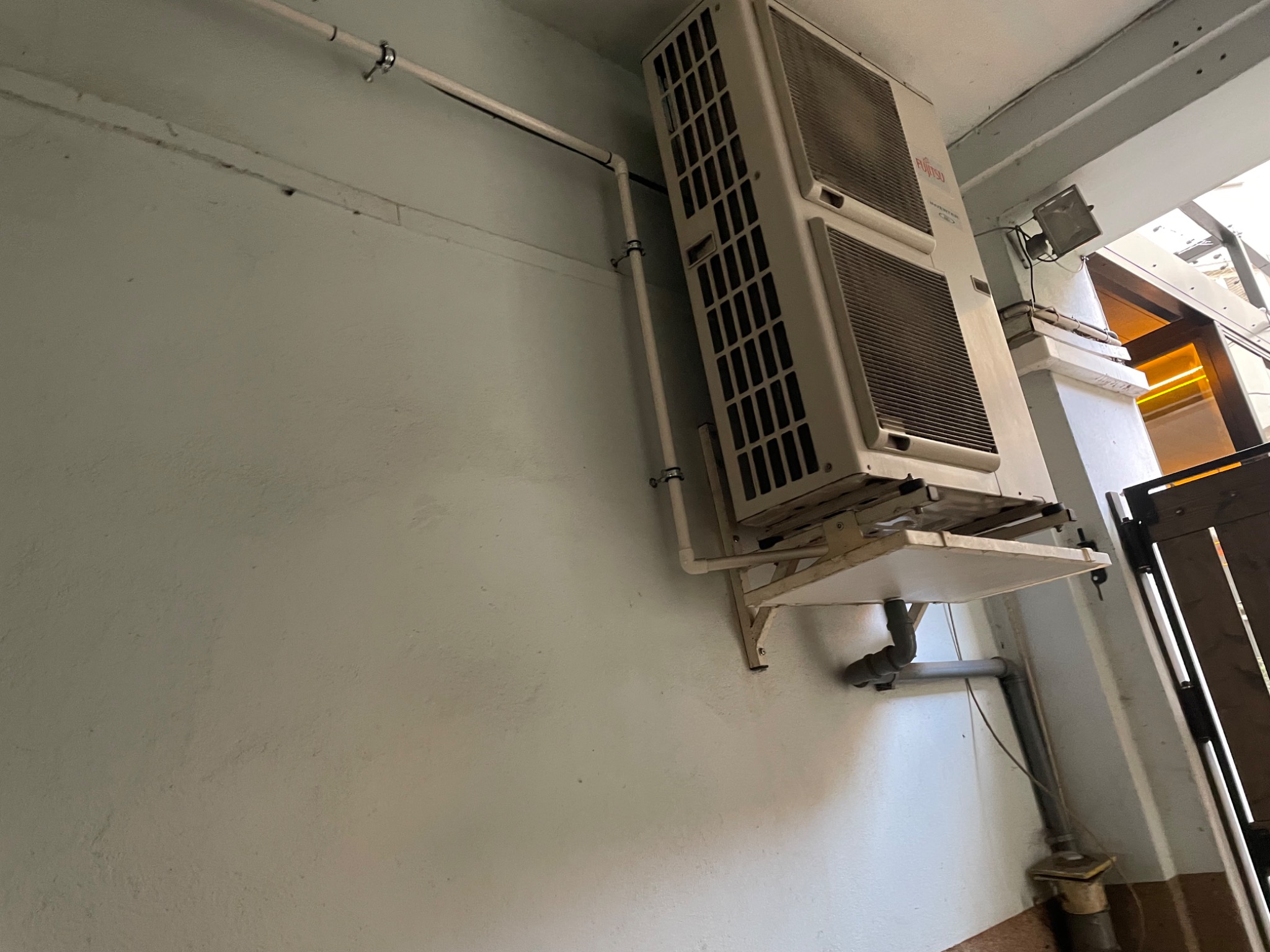 Biała, dwuczęściowa jednostka klimatyzacyjna Fujitsu zamontowana na zewnątrz budynku, z widocznymi rurami odprowadzającymi skropliny i przewodami elektrycznymi.