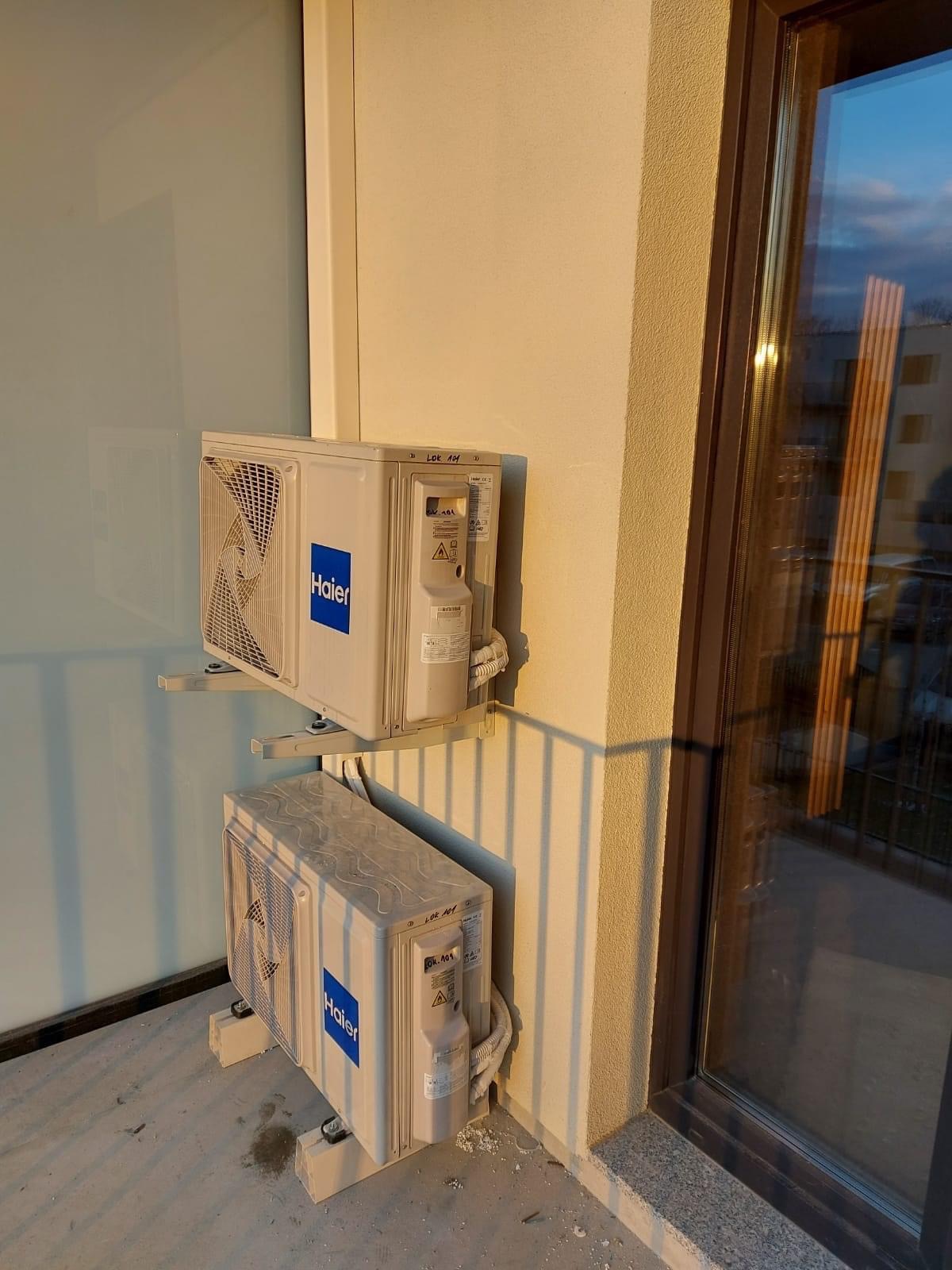 Dwa klimatyzatory Haier zamontowane na balkonie, jeden nad drugim, z widocznymi przewodami i uchwytami montażowymi, oświetlone popołudniowym słońcem.