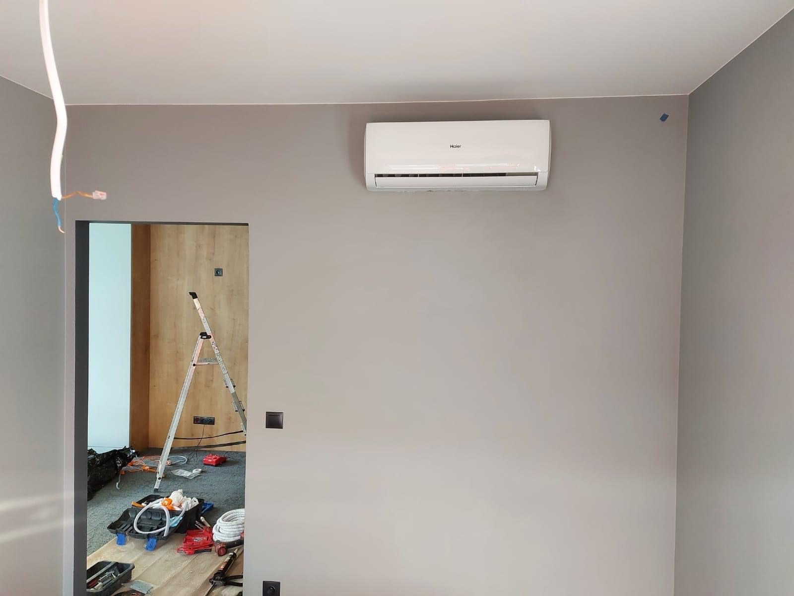 Biały klimatyzator Haier zamontowany na szarej ścianie w pokoju podczas instalacji, widoczne kable, drabina i narzędzia w tle.