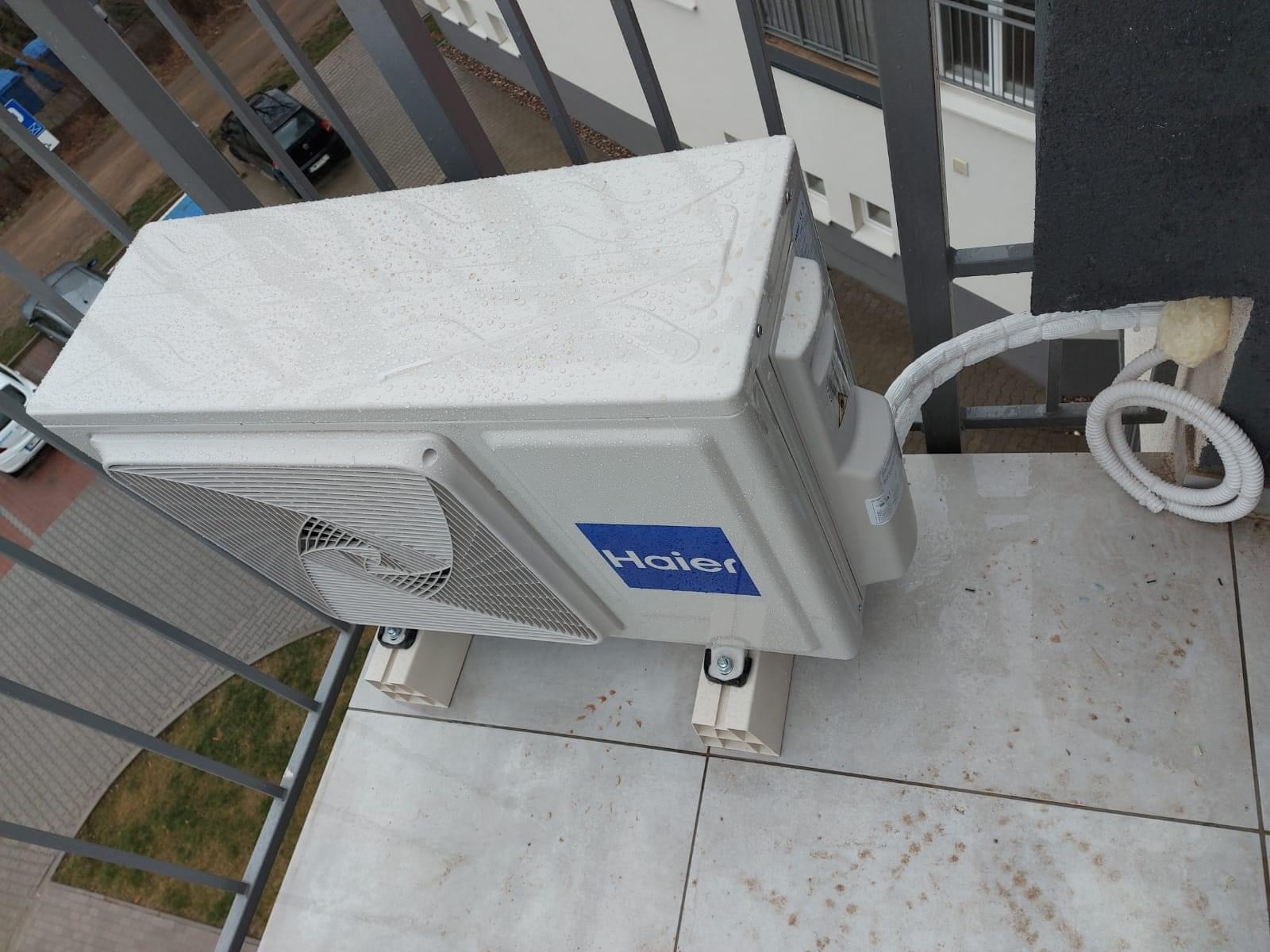 Biała jednostka zewnętrzna klimatyzatora Haier zamontowana na balkonie, widoczne krople wody na obudowie i izolowane rury przyłączeniowe.