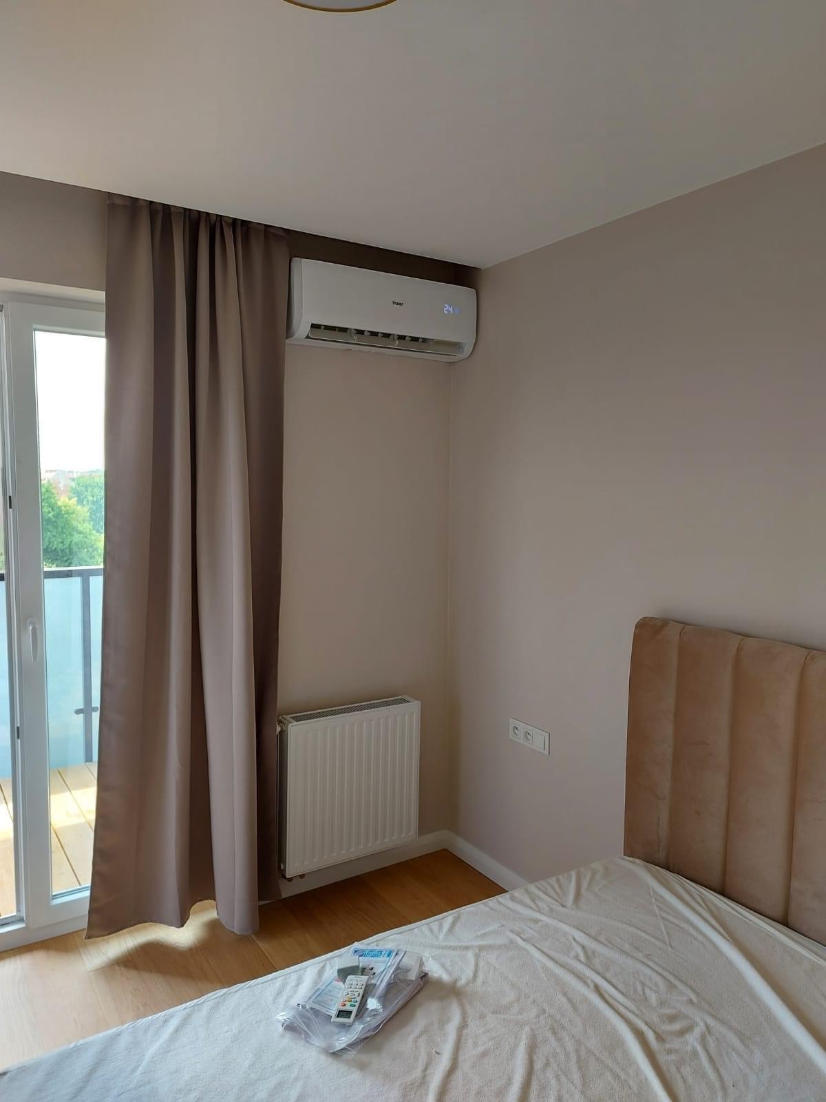 Biały klimatyzator Haier zamontowany nad grzejnikiem w sypialni, widok z boku łóżka, na którym leżą piloty i instrukcje.
