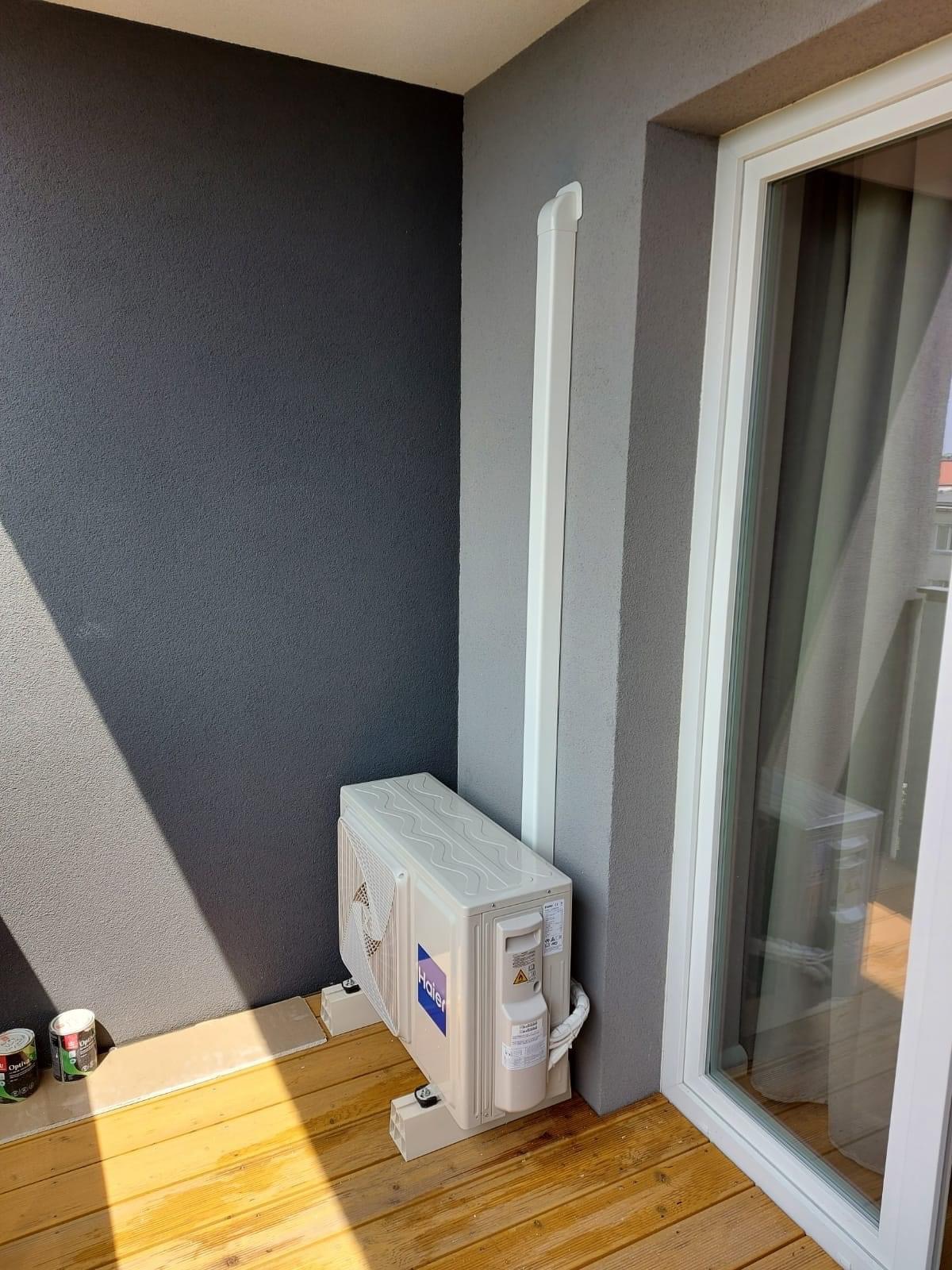 Zewnętrzna jednostka klimatyzacji Haier zamontowana na drewnianym balkonie, widoczne puszki z farbą, szara ściana i biała rura odprowadzająca skropliny.