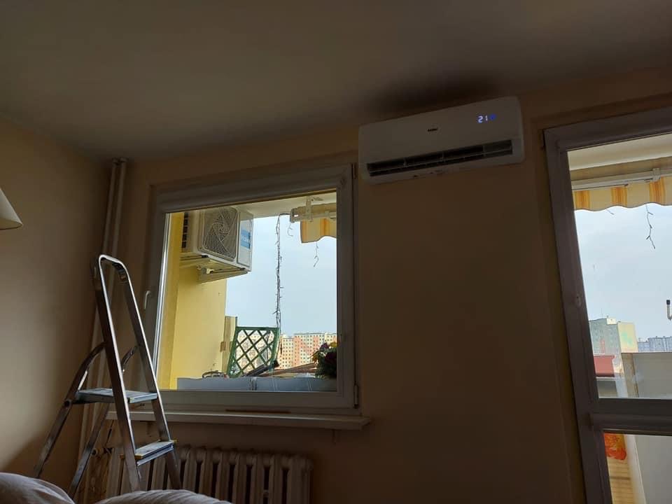 Biały klimatyzator Haier z wyświetlaczem temperatury 21 stopni zamontowany nad oknem w pokoju, obok stoi aluminiowa drabina, a za oknem widoczny balkon z jednostką zewnętrzną.