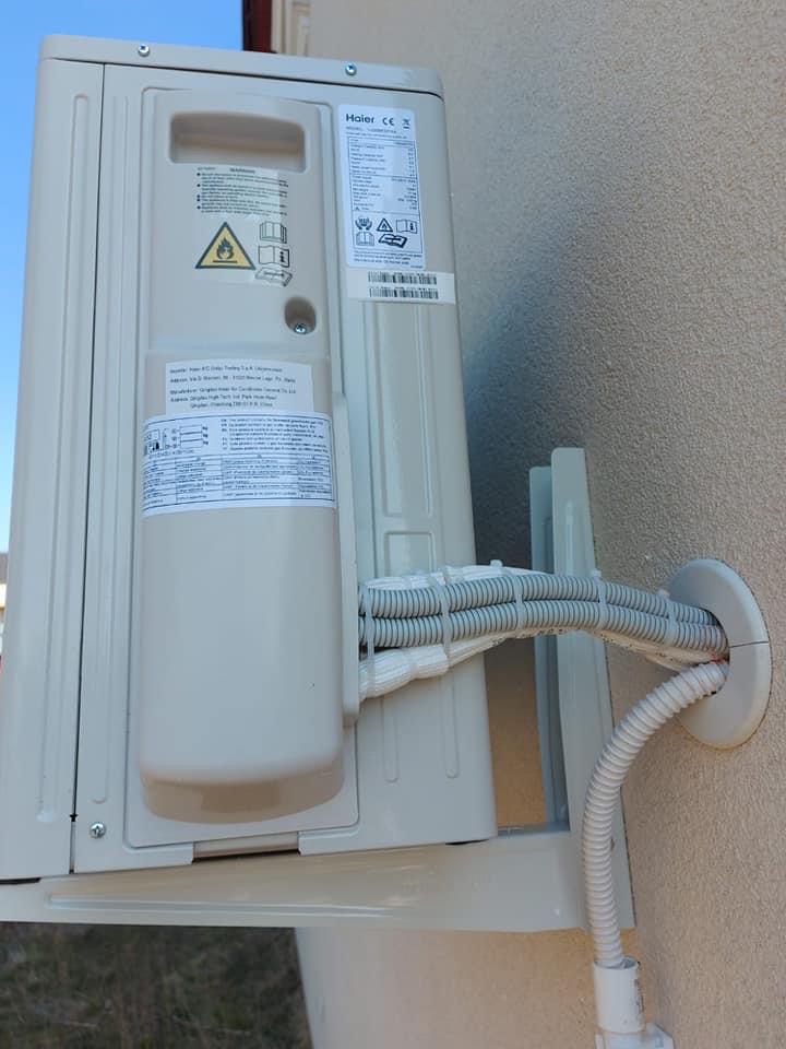 Zewnętrzna jednostka klimatyzacji Haier zamontowana na ścianie budynku, widoczne przewody w peszlach wchodzące przez otwór w ścianie.