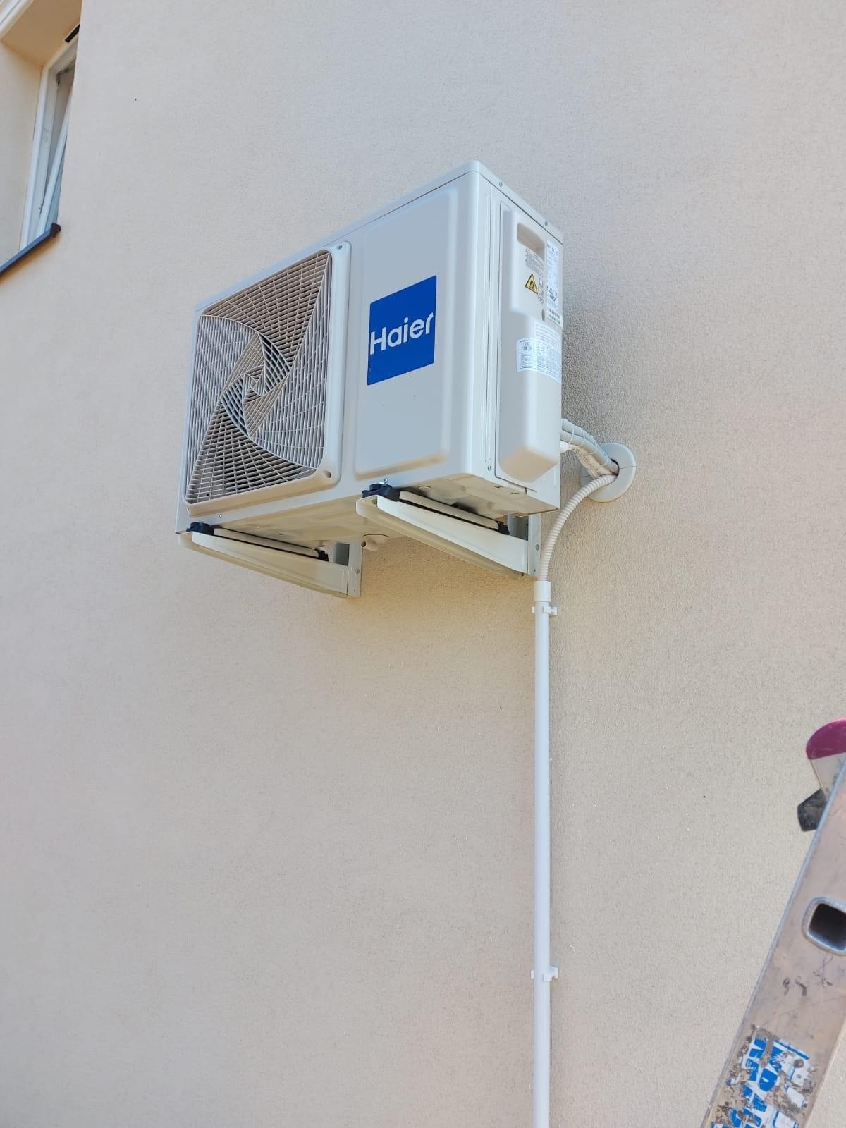 Biały klimatyzator Haier zamontowany na beżowej ścianie budynku, widoczna drabina aluminiowa z naklejkami.
