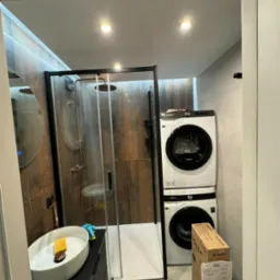 Wykończona łazienka z kabiną prysznicową typu walk-in z czarnymi ramami, umywalką nablatową na czarnej szafce z frezowanymi frontami oraz pralką i suszarką ustawionymi w pionie, dwie kartonowe...