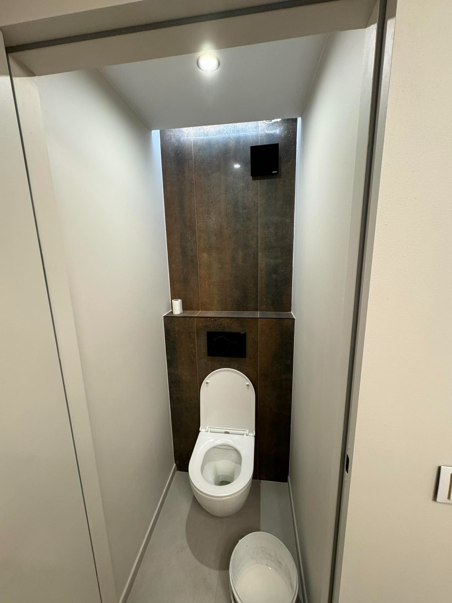 Wąska toaleta po remoncie z brązowymi płytkami na ścianie, białą miską ustępową i wiadrem z farbą w rogu. Podświetlony sufit i rolka papieru toaletowego.