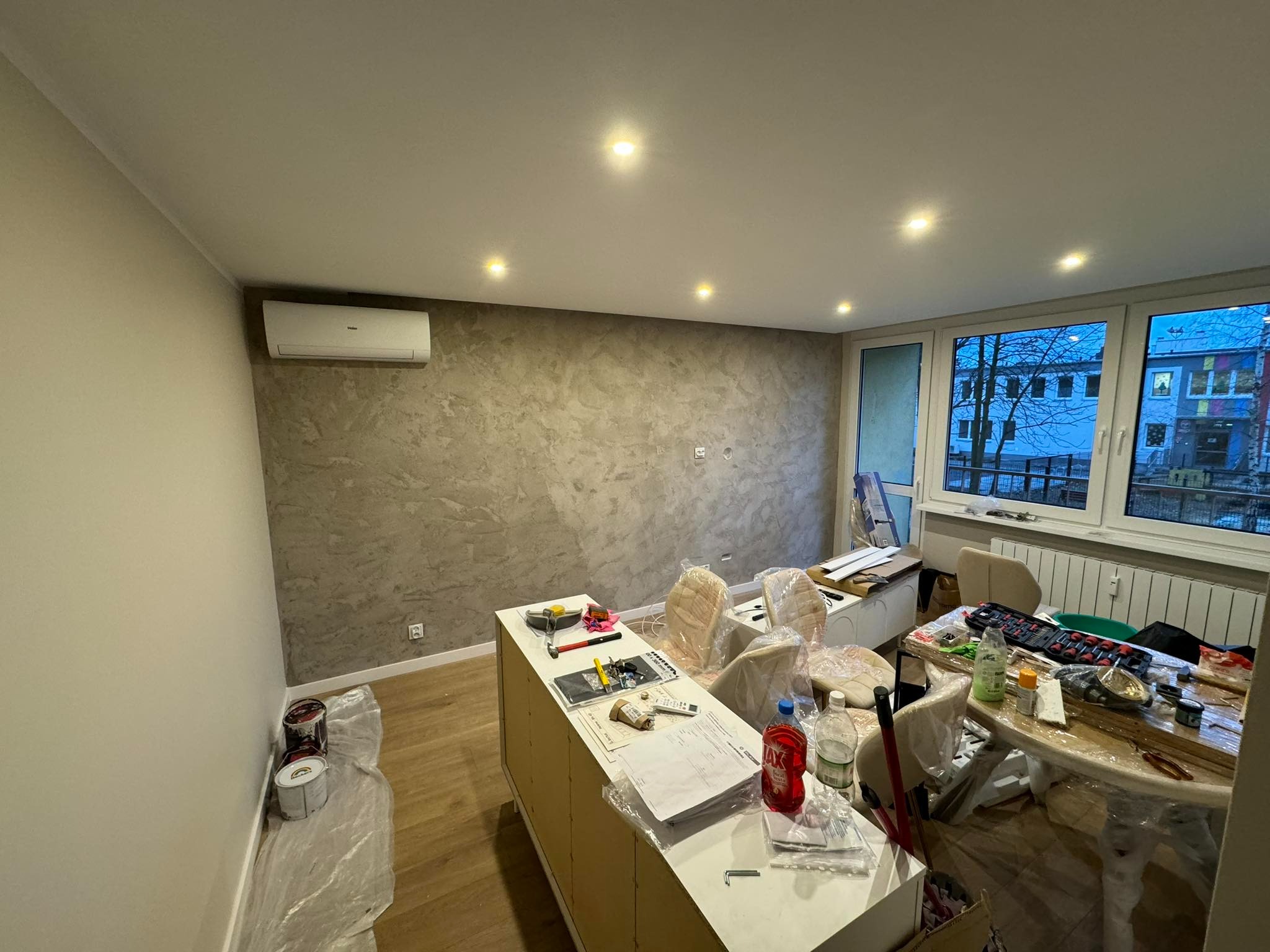 Pokój po remoncie z dekoracyjną szarą ścianą, widoczne puszki z farbą, narzędzia, zabezpieczone meble folią i okno z widokiem na budynek szkolny.