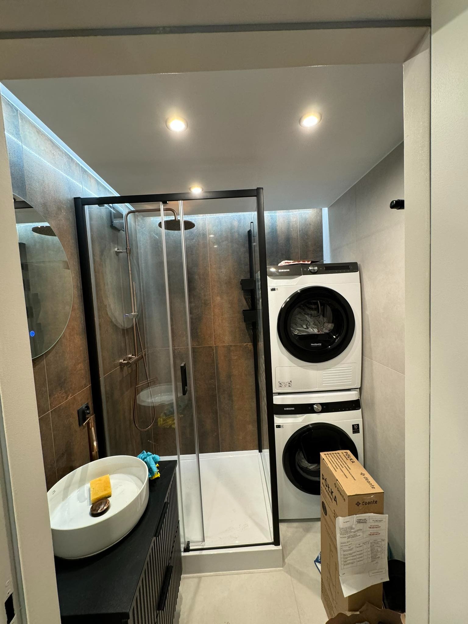 Wykończona łazienka z kabiną prysznicową typu walk-in z czarnymi ramami, umywalką nablatową na czarnej szafce z frezowanymi frontami oraz pralką i suszarką ustawionymi w pionie, dwie kartonowe...