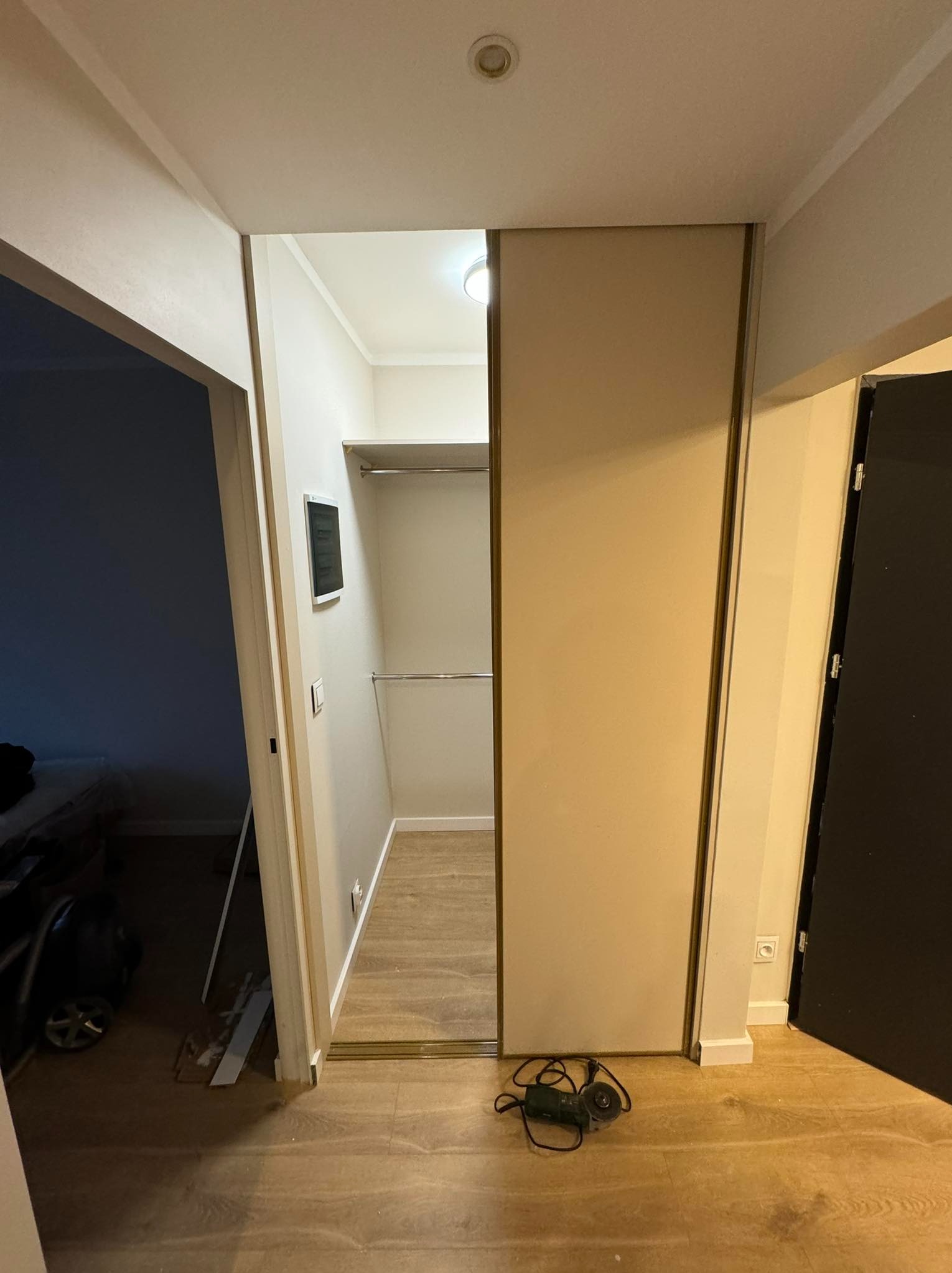 Widok na wnętrze garderoby z zamontowanymi przesuwnymi drzwiami, na podłodze leży szlifierka kątowa, w tle widoczny fragment sypialni z łóżkiem.