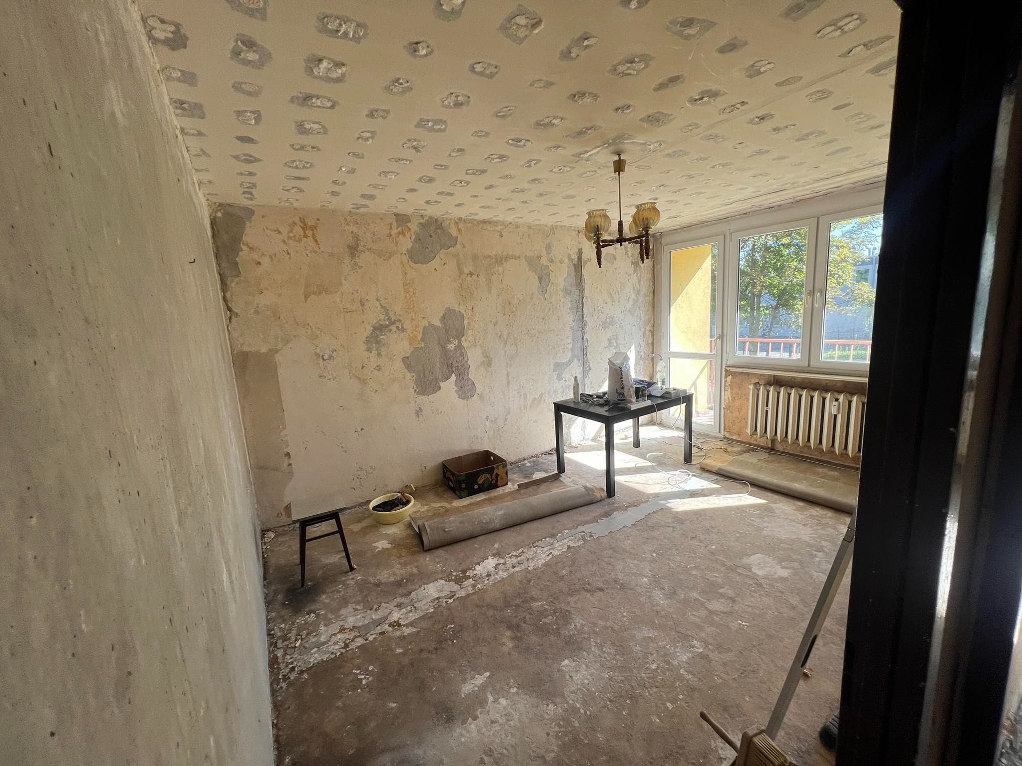 Pokój w trakcie remontu: ściany częściowo otynkowane, sufit z widocznymi łatami, stare okno z widokiem na drzewa, stolik z puszkami farb, zwinięty dywan, widoczny fragment grzejnika i drzwi.