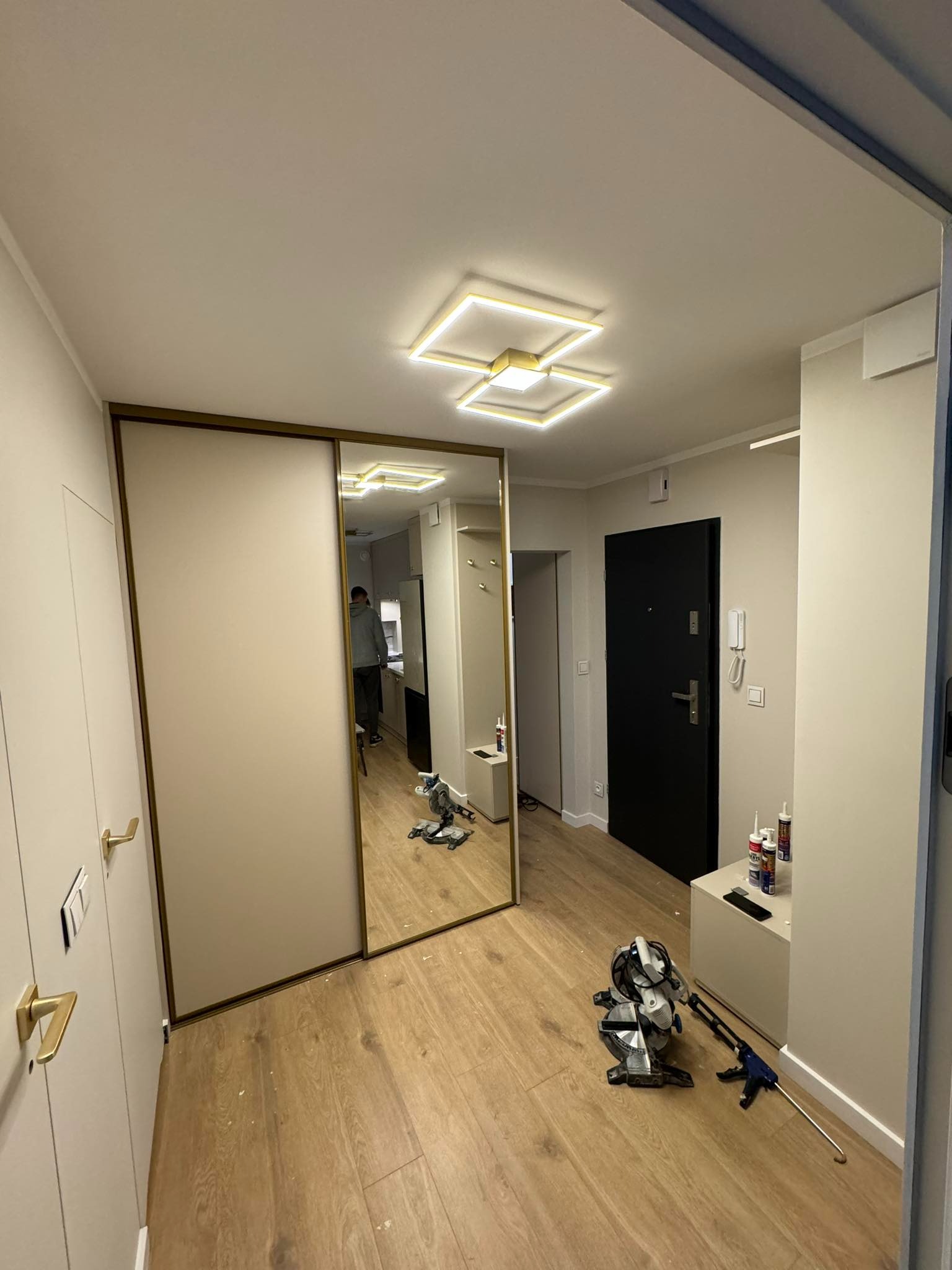 Korytarz po remoncie z przesuwną szafą z lustrem, drewnopodobną podłogą i nowoczesnym oświetleniem sufitowym. Na podłodze leży piła ukosowa i pistolet do silikonu, a na szafce stoją tubki z silikonem.