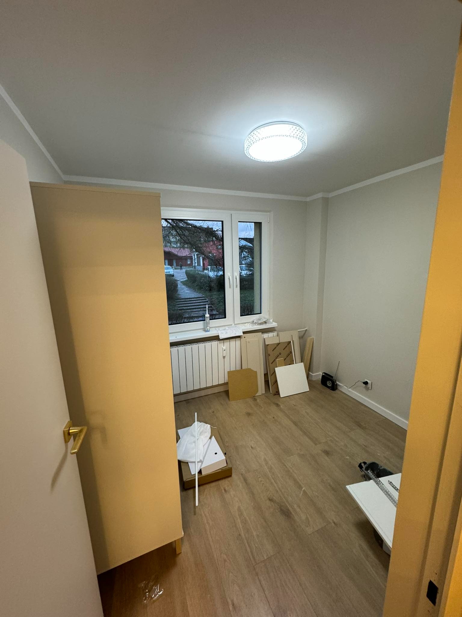 Pokój po odświeżeniu ścian, z nową podłogą i zamontowanym oknem; widoczne przygotowania do montażu drzwi, elementy ościeżnicy i drzwi oparte o ścianę.