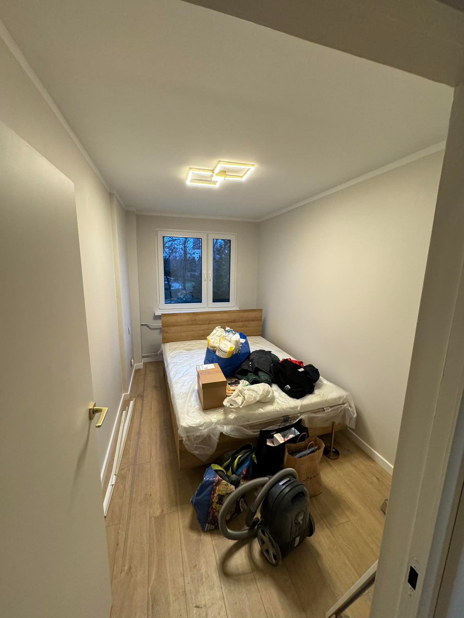 Pokój po remoncie z nowymi pomalowanymi ścianami w odcieniach beżu, drewnianą podłogą i sufitem z nowoczesną lampą, częściowo zagracony przedmioty i workami, z widocznym odkurzaczem.