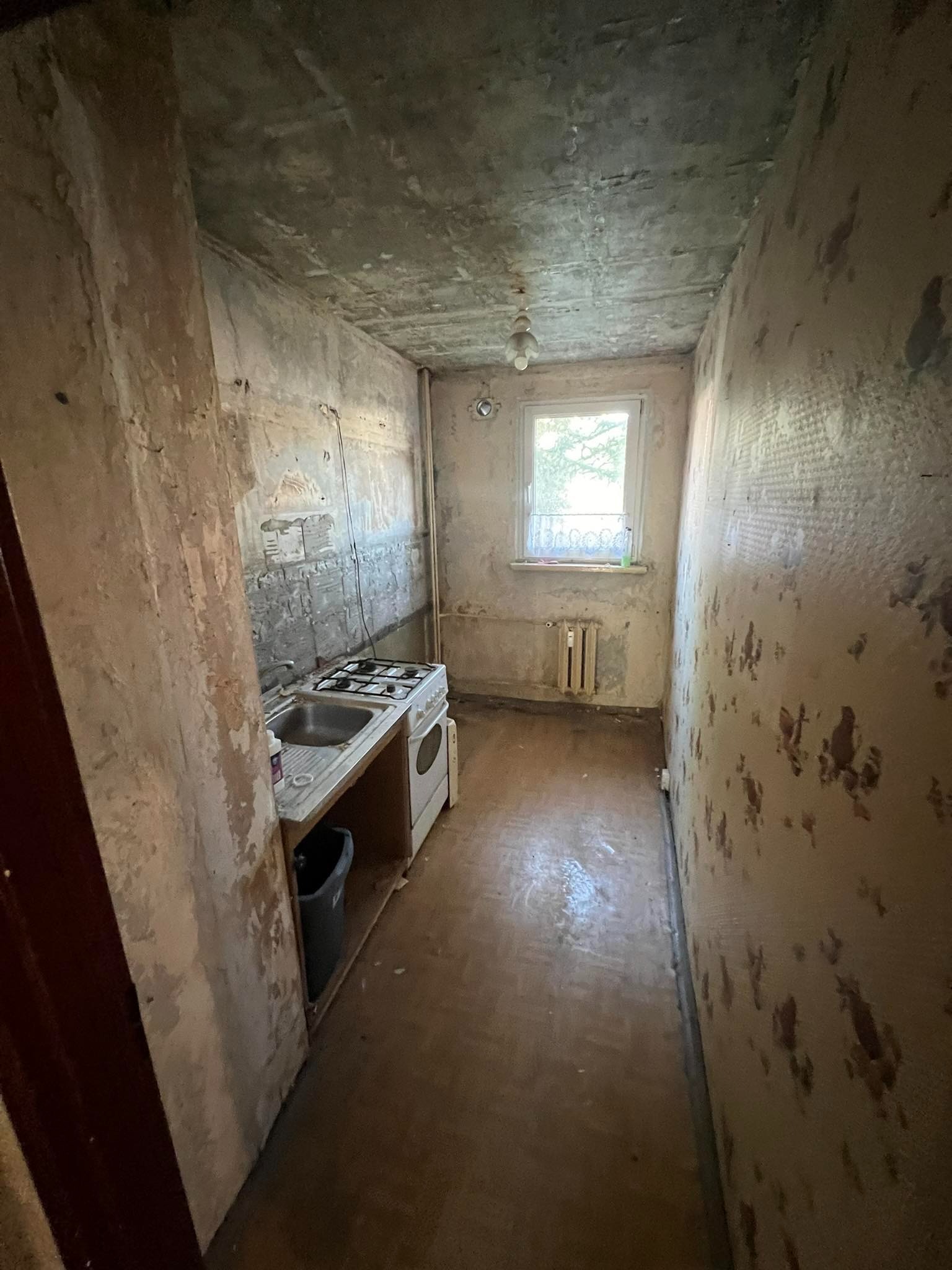 Wnętrze małej kuchni w stanie przed remontem: zniszczone ściany z odpadającą farbą i tapetą, stara kuchenka gazowa z zlewem, okno z firanką, podłoga z parkietu w złym stanie.