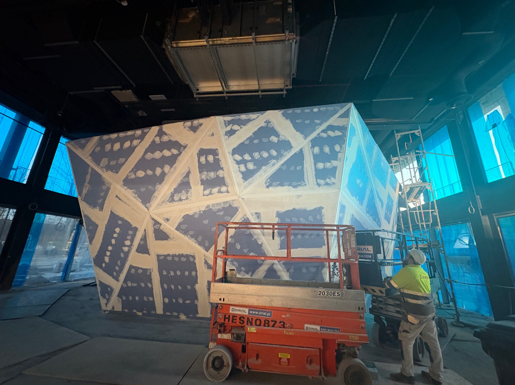 Szpachlowanie geometrycznej konstrukcji z płyt karton gips w Warszawie, widoczna podnośnik nożycowy i pracownik w odblaskowej kamizelce.