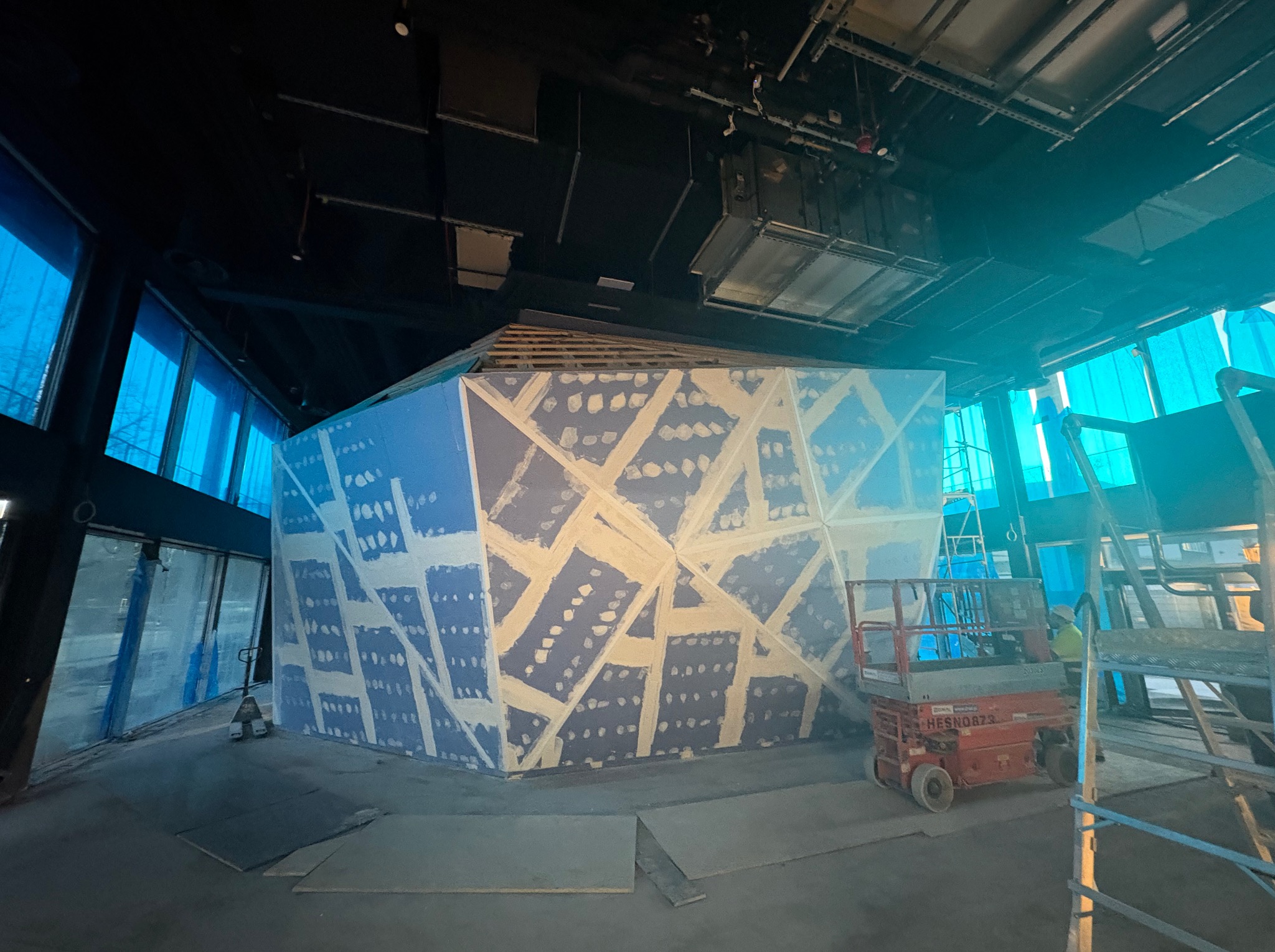 Wnętrze komercyjne w trakcie tworzenia konstrukcji z płyty kartonowo-gipsowej, widoczna szpachlowana siatka wzmacniająca na niebieskich płytach, podnośnik nożycowy w tle, surowy stan wykończenia.