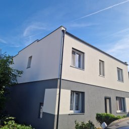 Eljot Elewacje PIOTR GRAJEWSKI - Dwukondygnacyjny dom z nowoczesną elewacją w kolorach szarości i bieli, widoczny z poziomu ogrodu w słoneczny dzień. Na niebie smugi kondensacyjne samolotów.