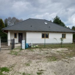 Eljot Elewacje PIOTR GRAJEWSKI - Parterowy dom z jasną elewacją i grafitowym dachem, widoczne ślady prac budowlanych, ogrodzenie z siatki, ziemia z fragmentami trawy, mężczyzna w drzwiach.