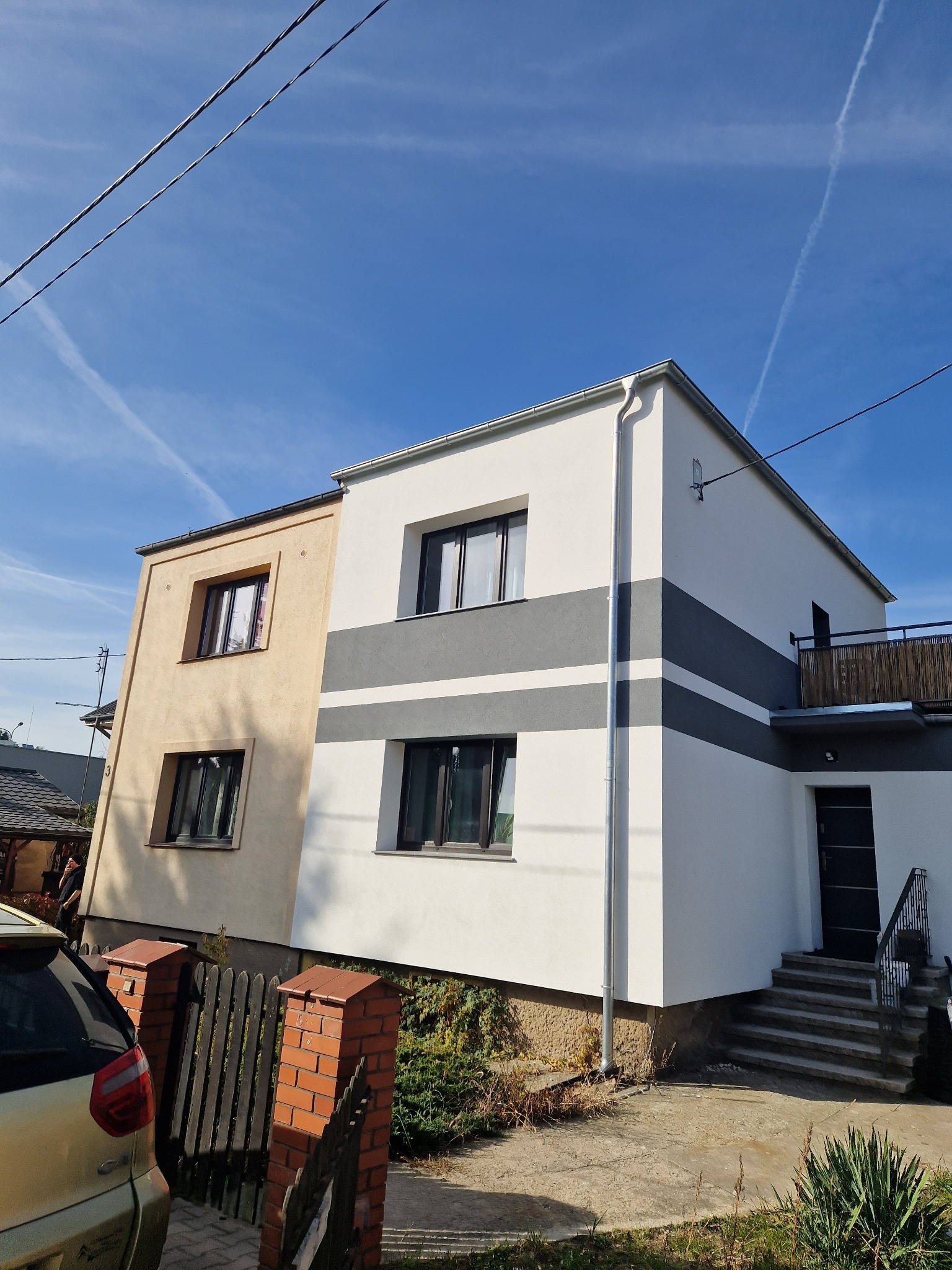 Dwukondygnacyjny dom z elewacją w dwóch odcieniach: beżowym i białym z szarymi pasami, widoczny z zewnątrz, z samochodem na pierwszym planie i błękitnym niebem w tle.