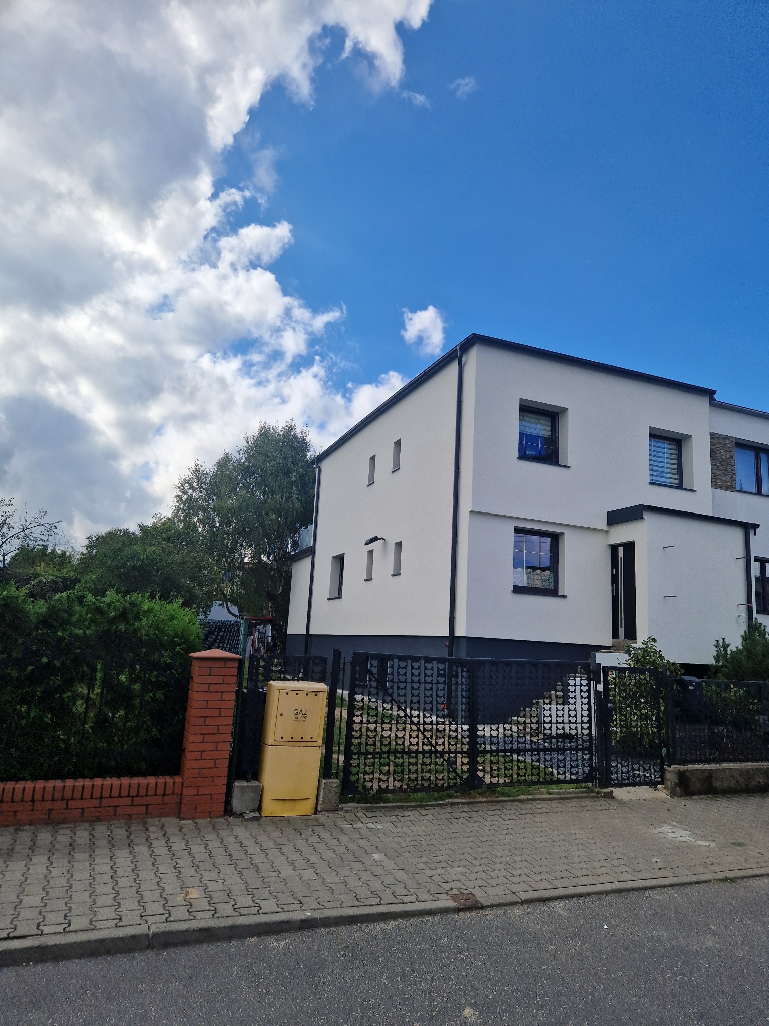 Nowoczesny, dwukondygnacyjny dom z białą elewacją i czarnymi akcentami, widoczny z poziomu ulicy, z ozdobnym metalowym ogrodzeniem i skrzynką gazową.