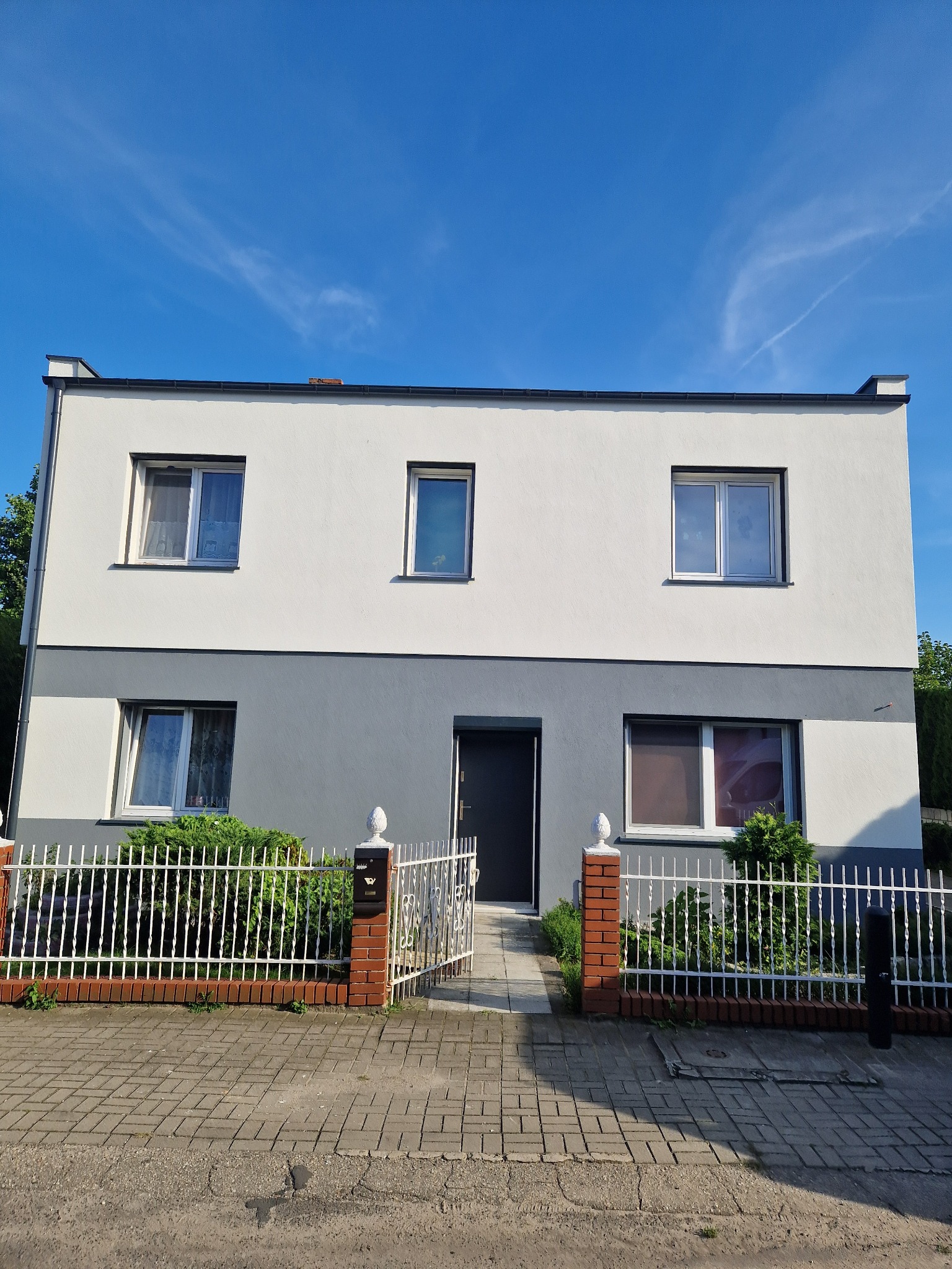 Dwukondygnacyjny dom z biało-szarą elewacją, ogrodzony białym płotem z ceglanymi słupkami, widziany w słoneczny dzień.