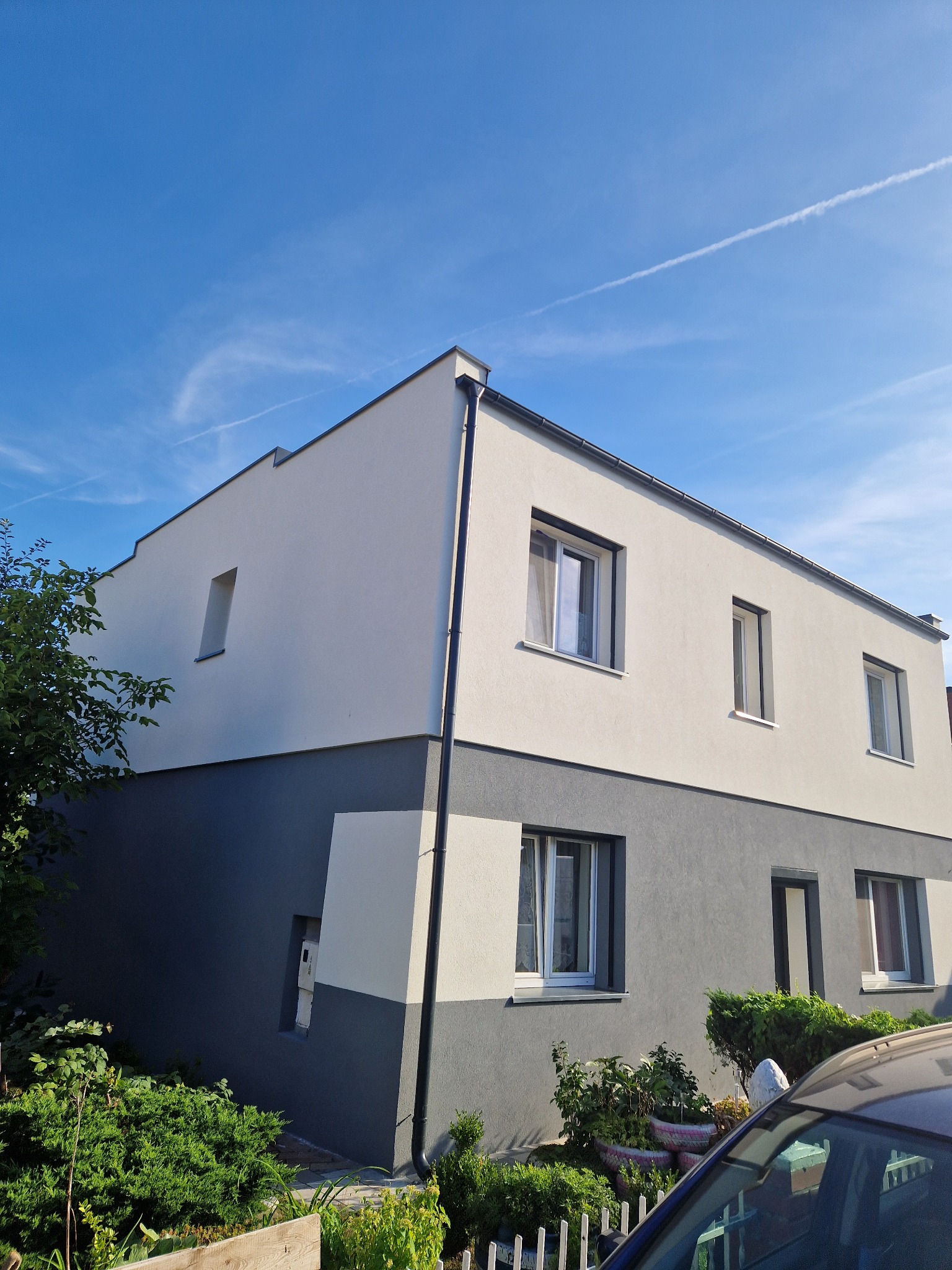 Dwukondygnacyjny dom z nowoczesną elewacją w kolorach szarości i bieli, widoczny z poziomu ogrodu w słoneczny dzień. Na niebie smugi kondensacyjne samolotów.