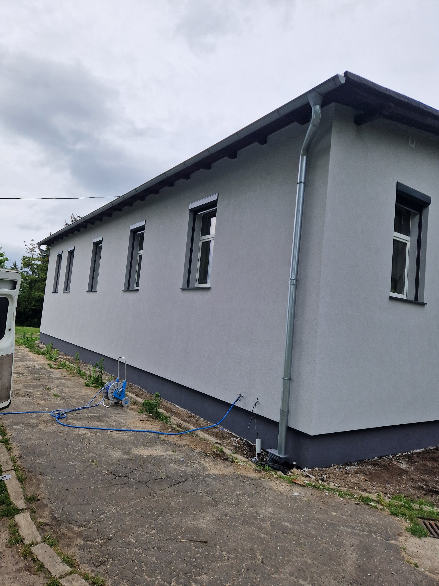 Świeżo otynkowana, szara elewacja budynku z widocznym systemem rynnowym, oknami z roletami zewnętrznymi i wężem do mycia pod ciśnieniem na chodniku.