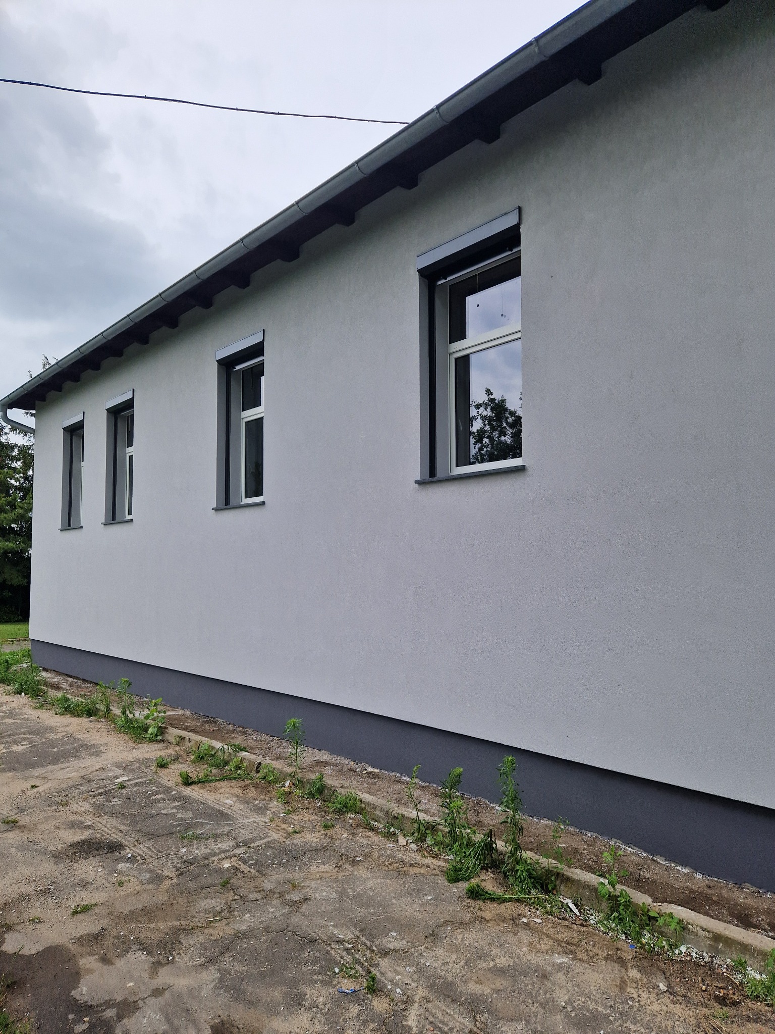 Świeżo otynkowana, szara elewacja budynku z trzema oknami z szarymi ramami i roletami, widoczne ślady po pracach budowlanych na ziemi.