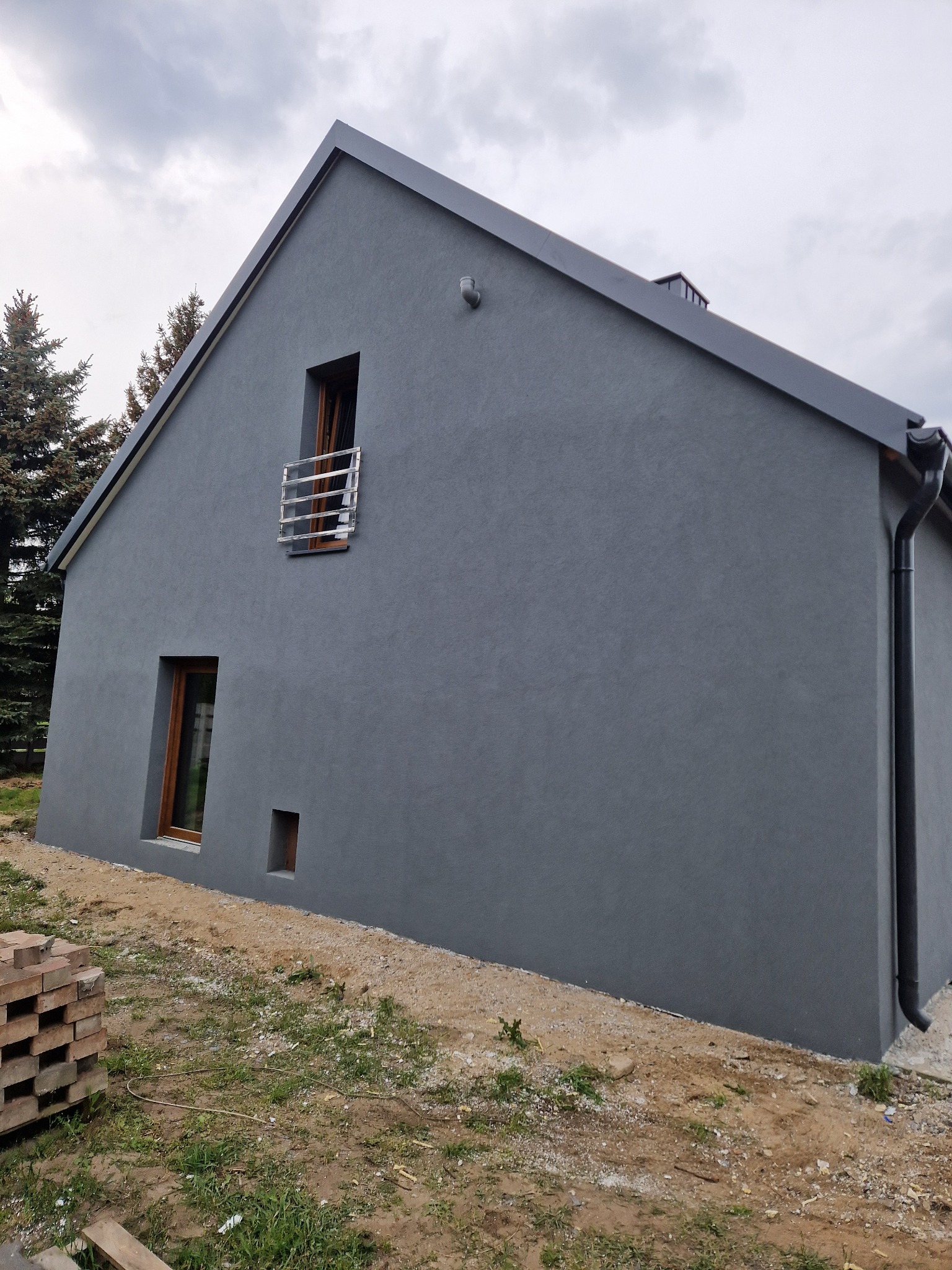 Szara elewacja domu jednorodzinnego z drewnianymi oknami i metalową balustradą, widoczne cegły na ziemi sugerują prace wykończeniowe.