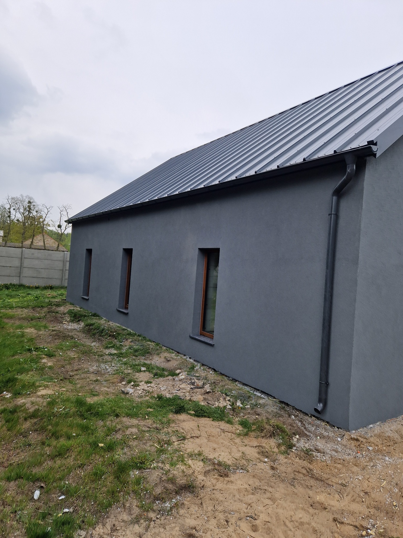 Szary dom jednorodzinny z blachy trapezowej na dachu, widok z boku z widocznymi trzema oknami i rynną spustową, na pierwszym planie trawa i piasek.