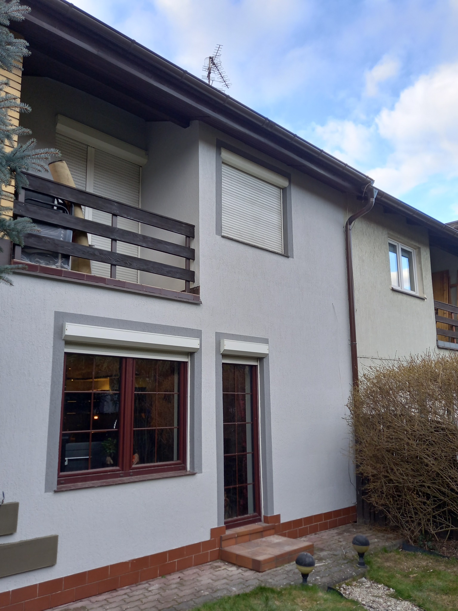 Dwukondygnacyjny dom z szarą elewacją, brązowymi ramami okiennymi i roletami, widok z zewnątrz na balkon i wejście, z elementami zieleni w otoczeniu.