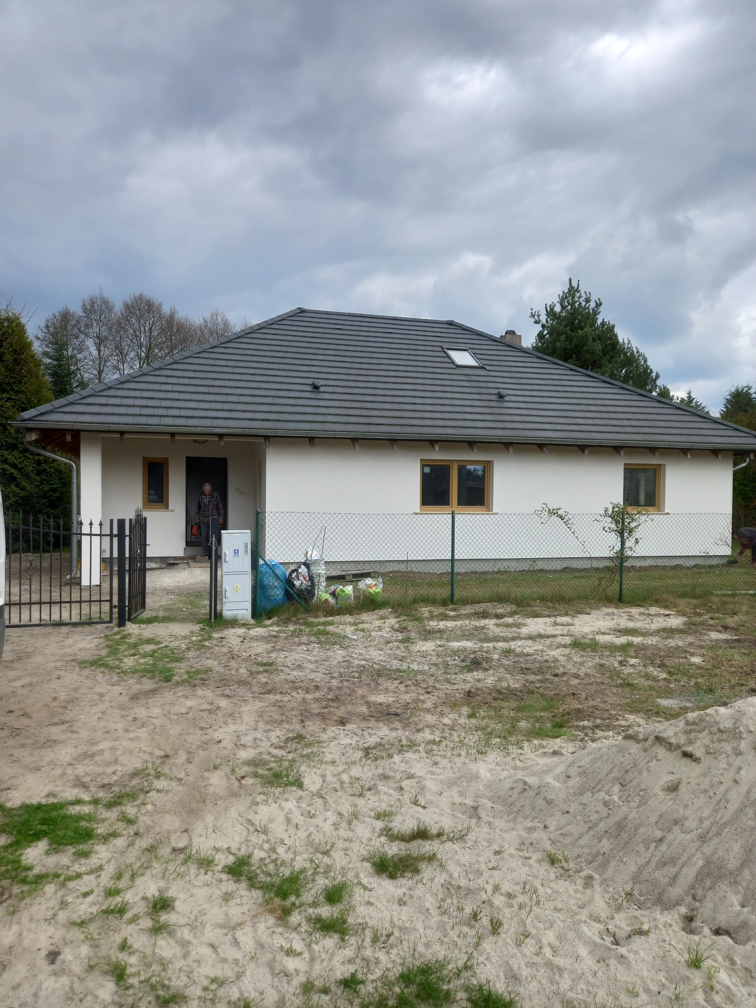 Parterowy dom z jasną elewacją i grafitowym dachem, widoczne ślady prac budowlanych, ogrodzenie z siatki, ziemia z fragmentami trawy, mężczyzna w drzwiach.
