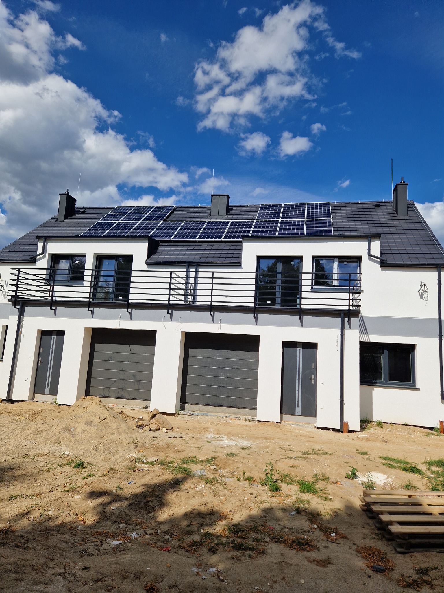 Bliźniak w nowoczesnym stylu z panelami słonecznymi na dachu, czarnymi ramami okiennymi i balkonami z metalowymi barierkami, otoczony piaszczystym terenem.