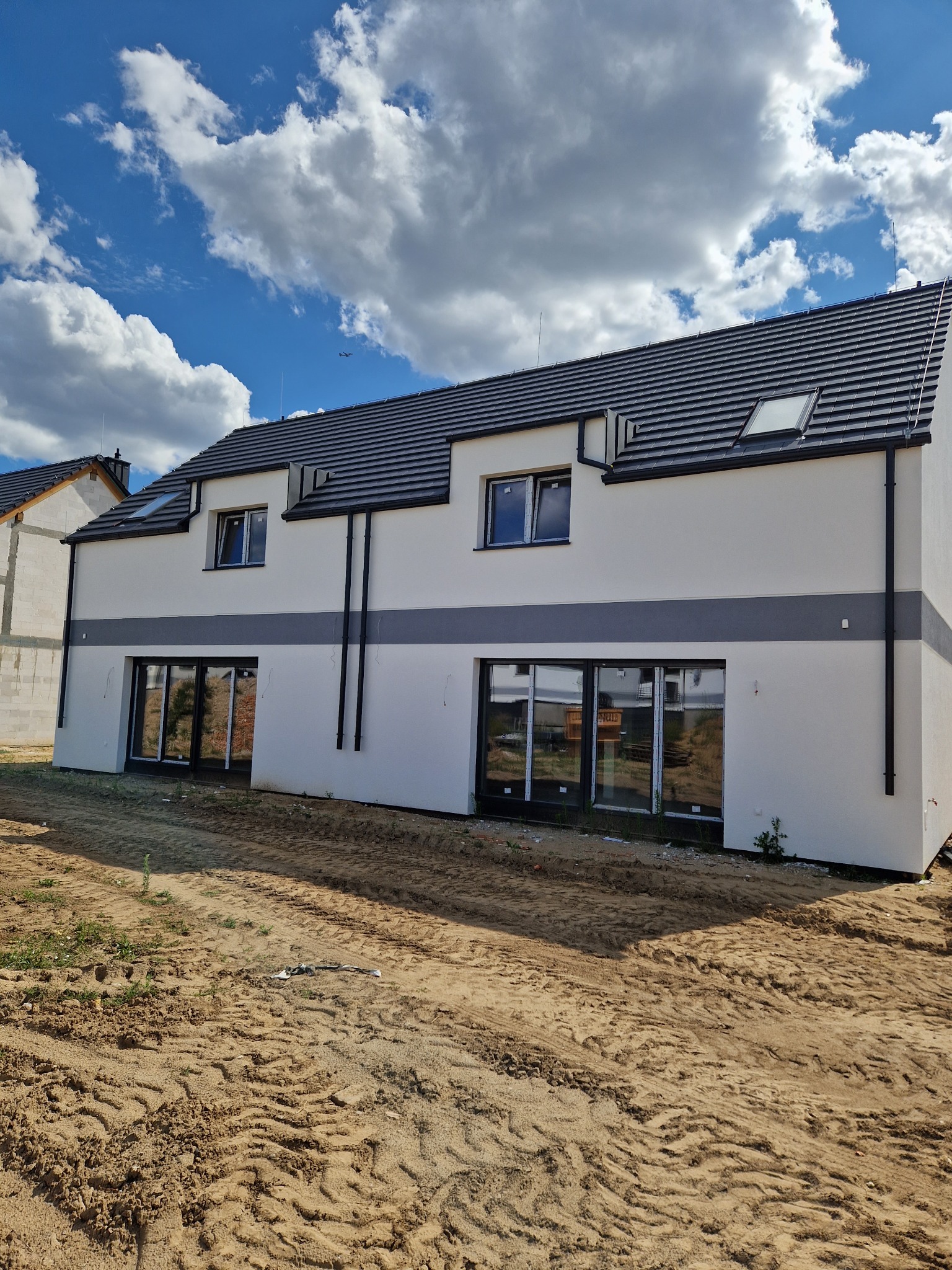 Nowoczesny dom dwukondygnacyjny z białą elewacją i szarym pasem dekoracyjnym, czarną dachówką i oknami, na tle błękitnego nieba z obłokami, teren wokół domu w stanie surowym.