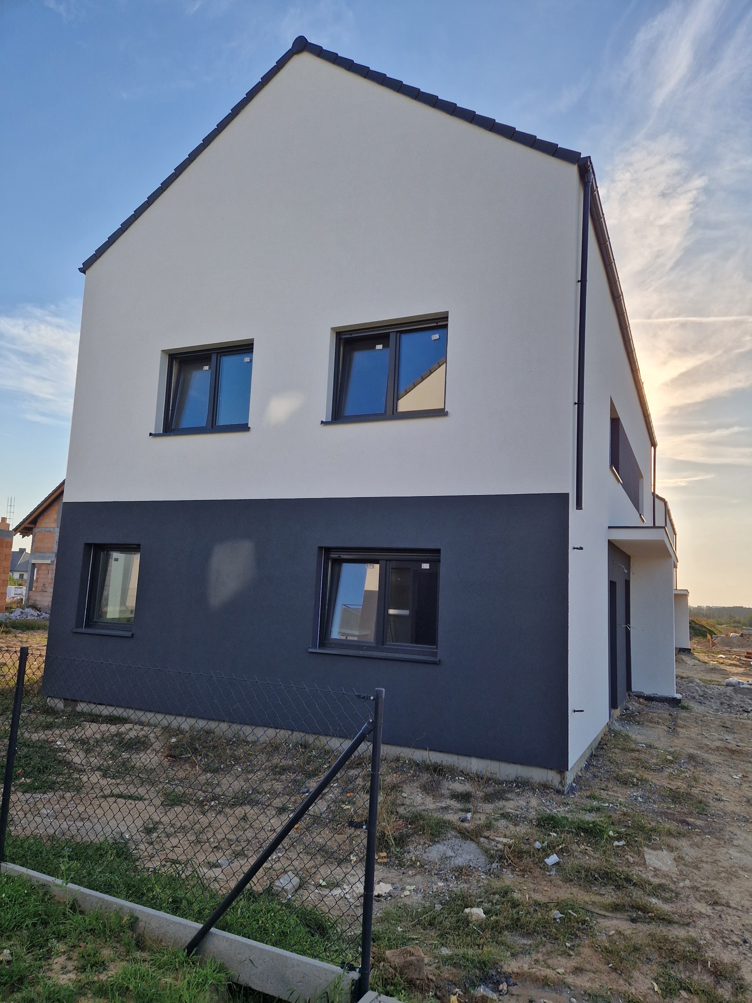 Dwukondygnacyjny dom z elewacją w kolorze białym i grafitowym, czarne ramy okienne, widok z zewnątrz na tle błękitnego nieba z chmurami.