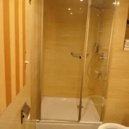 Wykończona łazienka z kabiną prysznicową z przezroczystego szkła, biały brodzik, toaleta w kolorze ecru, ściany wyłożone płytkami w odcieniach beżu i brązu, pionowe pasy na ścianie po lewej stronie.