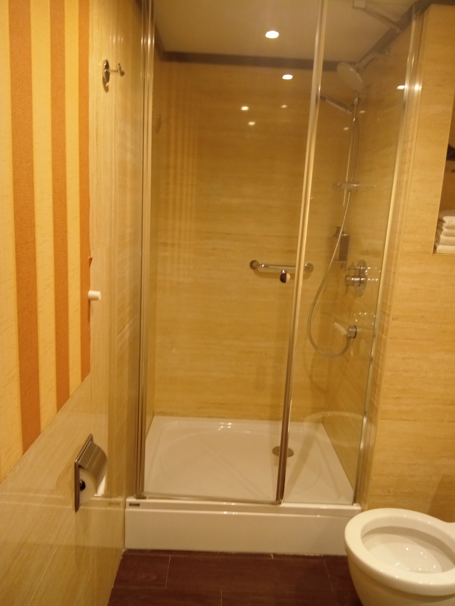 Wykończona łazienka z kabiną prysznicową z przezroczystego szkła, biały brodzik, toaleta w kolorze ecru, ściany wyłożone płytkami w odcieniach beżu i brązu, pionowe pasy na ścianie po lewej stronie.