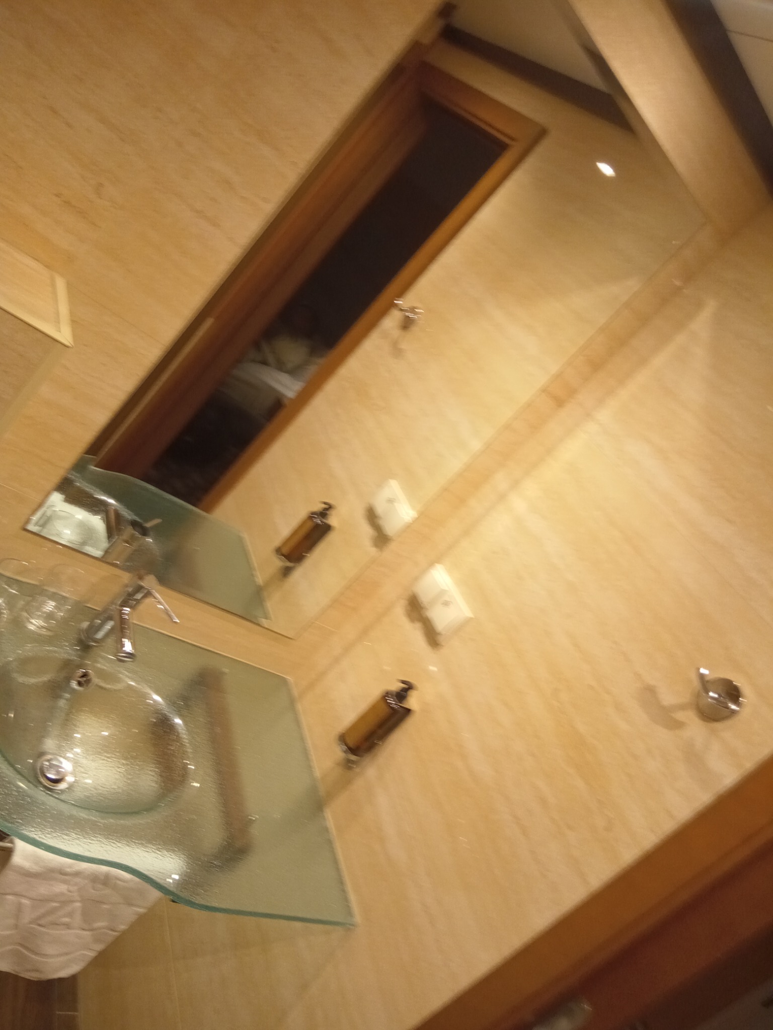 Umywalka ze szklanym blatem w łazience wyłożonej płytkami imitującymi drewno, widoczne dozowniki mydła i lustro odbijające wnętrze